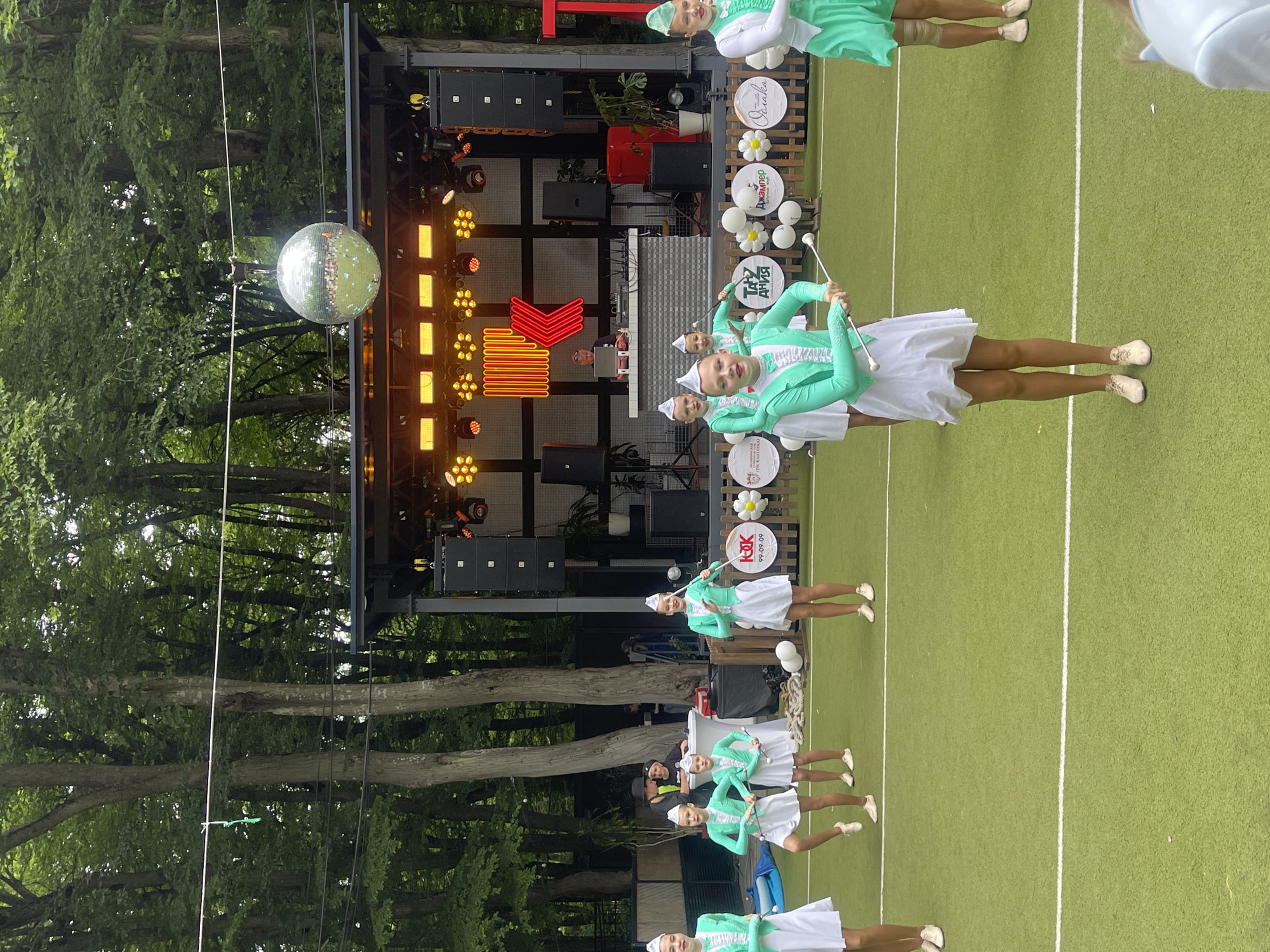 In Stavropol, Russia, a group of young women in matching teal and white uniforms are performing a baton twirling routine on a green artificial turf field. They are positioned in front of a stage with a DJ booth and a large disco ball hanging above. The background consists of a dense forest of trees, suggesting an outdoor event or park setting. The women are holding batons, some are in motion, and their expressions appear focused and engaged in their performance. The event seems to be taking place during the day, under an overcast sky.