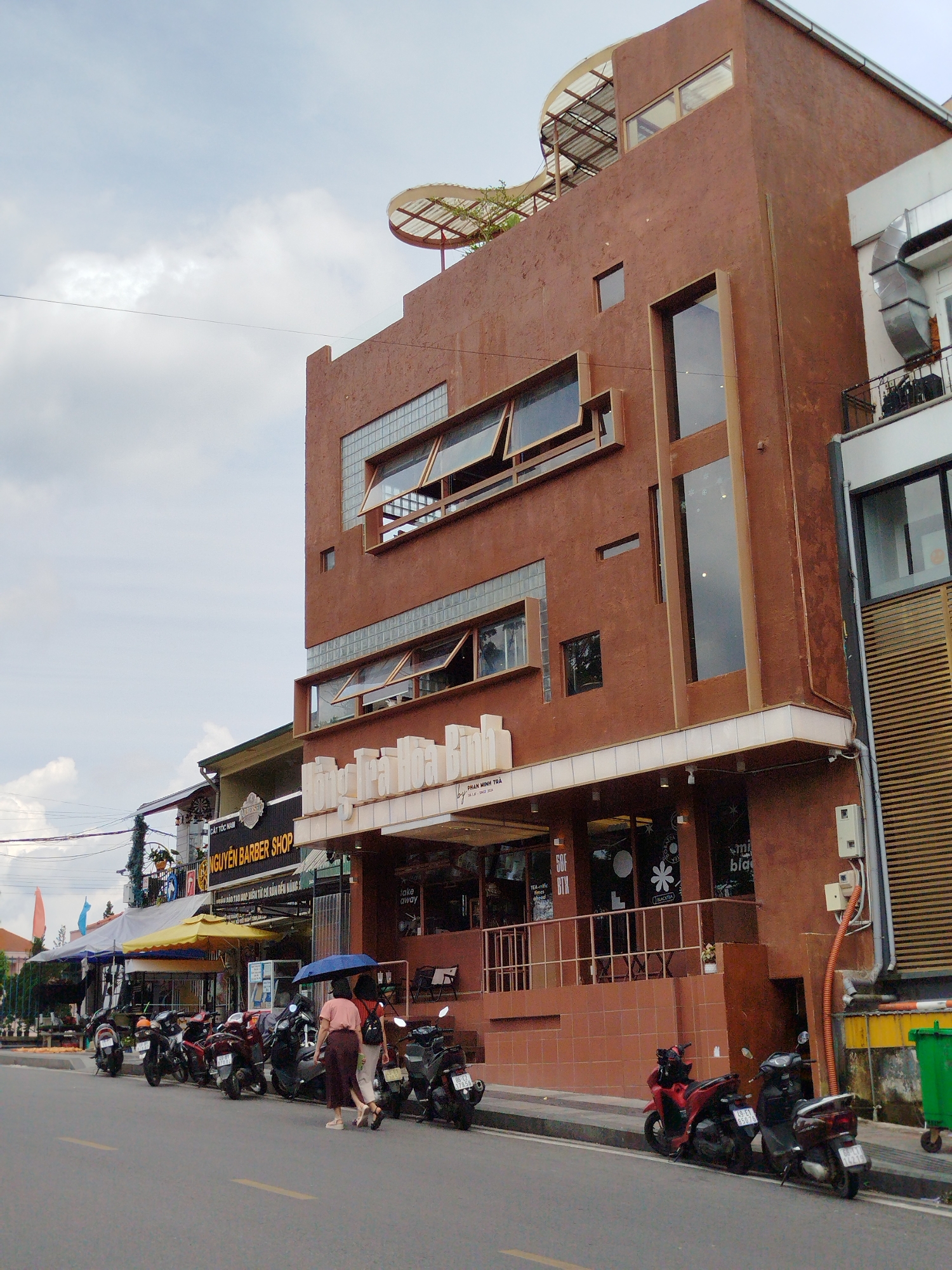 The image shows a street scene in Da Lat, Vietnam, featuring a prominent, reddish-brown, multi-story building on the right. The building has a distinctive architectural style with large, angled windows and a curved, translucent canopy on the rooftop. Several motorcycles are parked along the sidewalk in front of various businesses, including a "NGUYEN BARBER SHOP." Two women, one wearing a pink top and the other a white shirt, are walking on the sidewalk, one holding a blue umbrella. The sky is cloudy. The street is paved, and a yellow line marks the center of the road. To the far right, a green trash bin is visible. The overall scene depicts a typical urban street in Da Lat with its characteristic architecture and prevalent use of motorcycles.