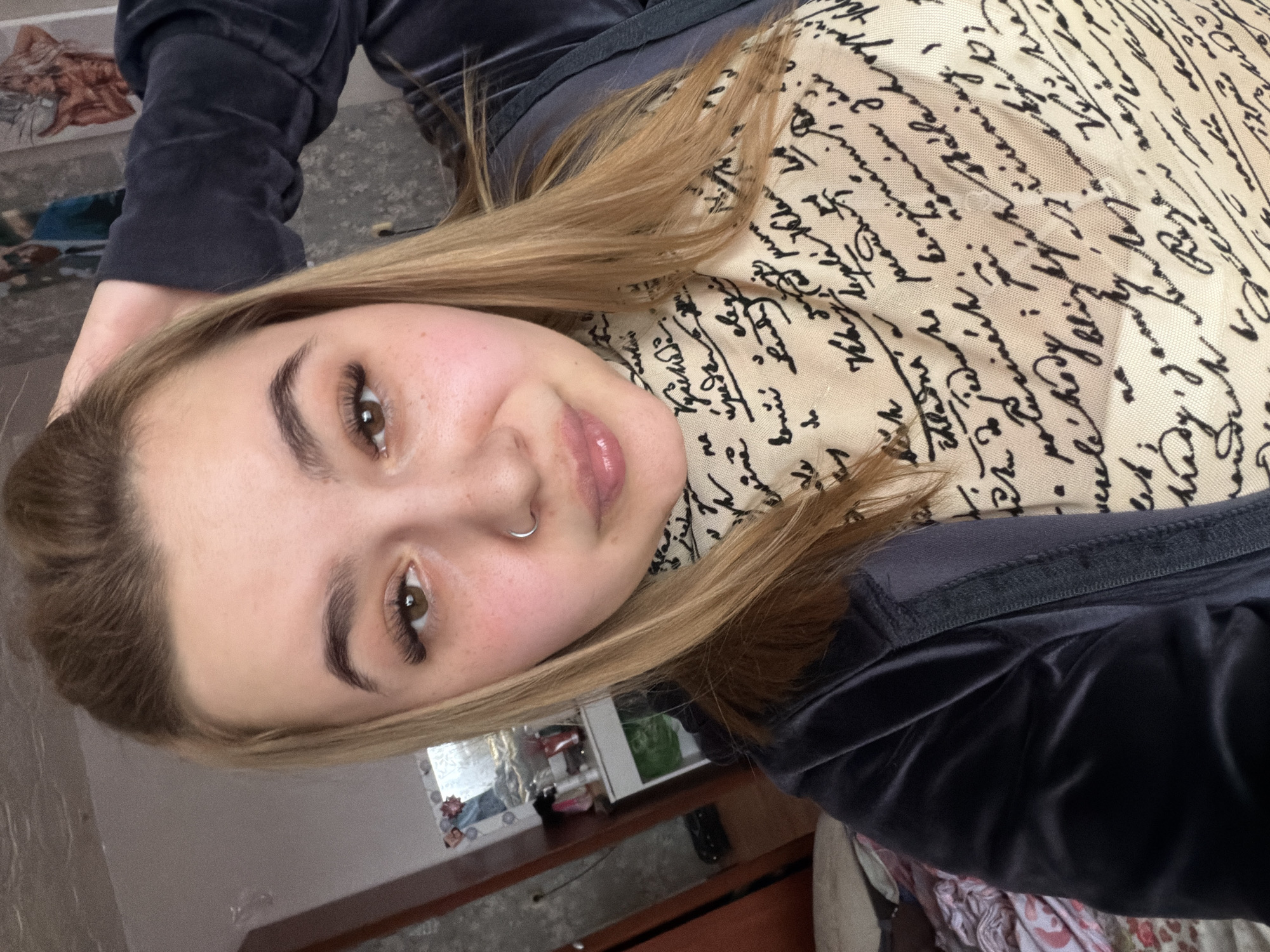 A close-up, slightly angled shot shows a young woman with light brown hair, brown eyes, and a nose ring. She is wearing a dark blue velvet tracksuit with a scarf or top that has a cream base printed with black cursive text. The background is blurred, revealing a room with a dresser and decorative items. The text on the scarf appears to be illegible handwriting. The image provides no information to suggest the location is Chaikovsky, Russia, or any specific location.