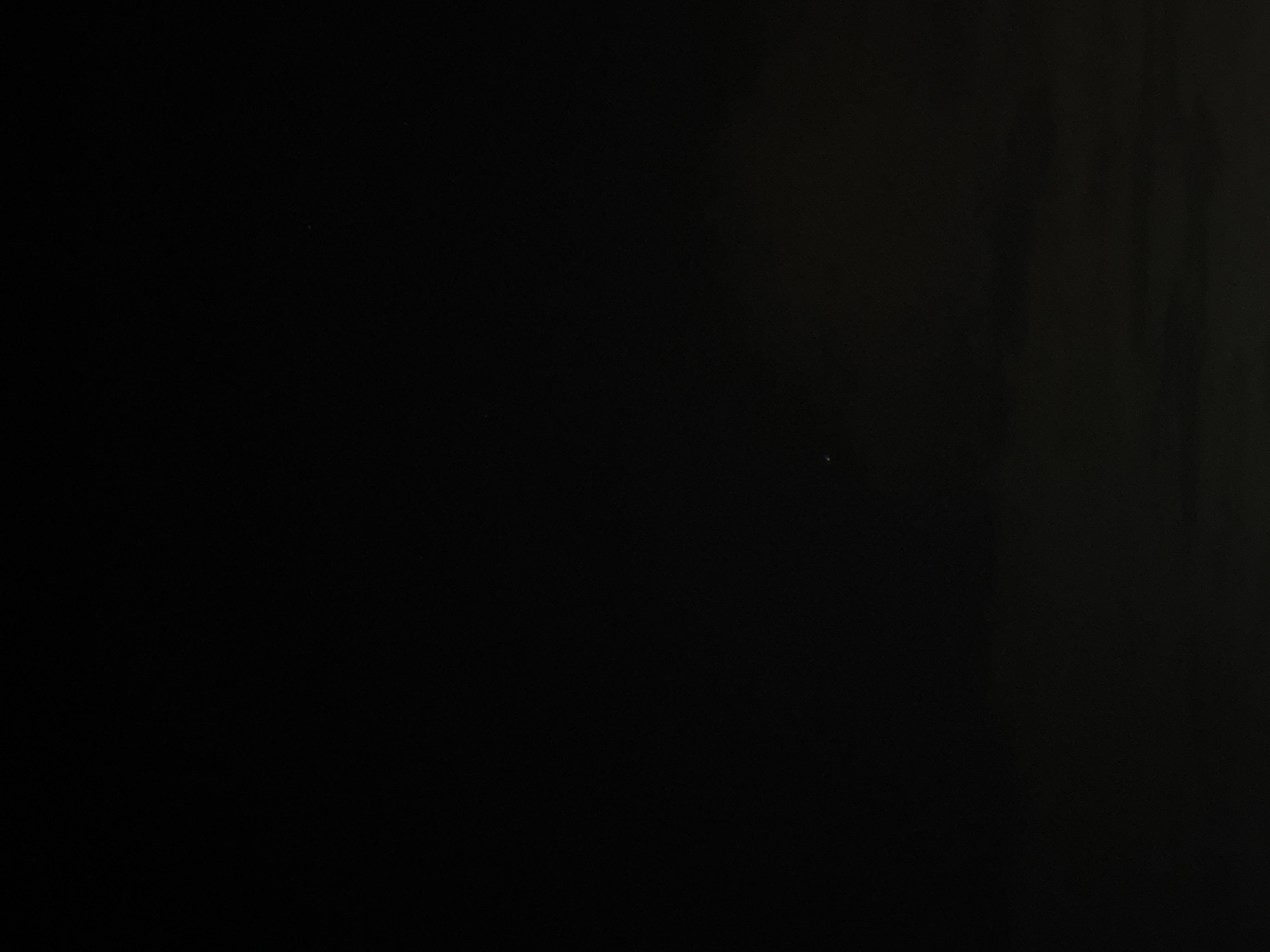 The image is a very dark, almost entirely black photograph, likely taken at night. There are faint, subtle horizontal bands of lighter gray in the lower portion of the image, suggesting a landscape or perhaps distant lights. The only distinct element is a single, small, white speck of light located in the lower-middle section of the frame, which could be a star or a distant light source. The image is extremely underexposed, making it difficult to discern any specific details of the environment. The provided crops offer no additional clarity or features. Without further context, it is impossible to determine if this image was taken in Nha Trang, Vietnam, or any specific location.