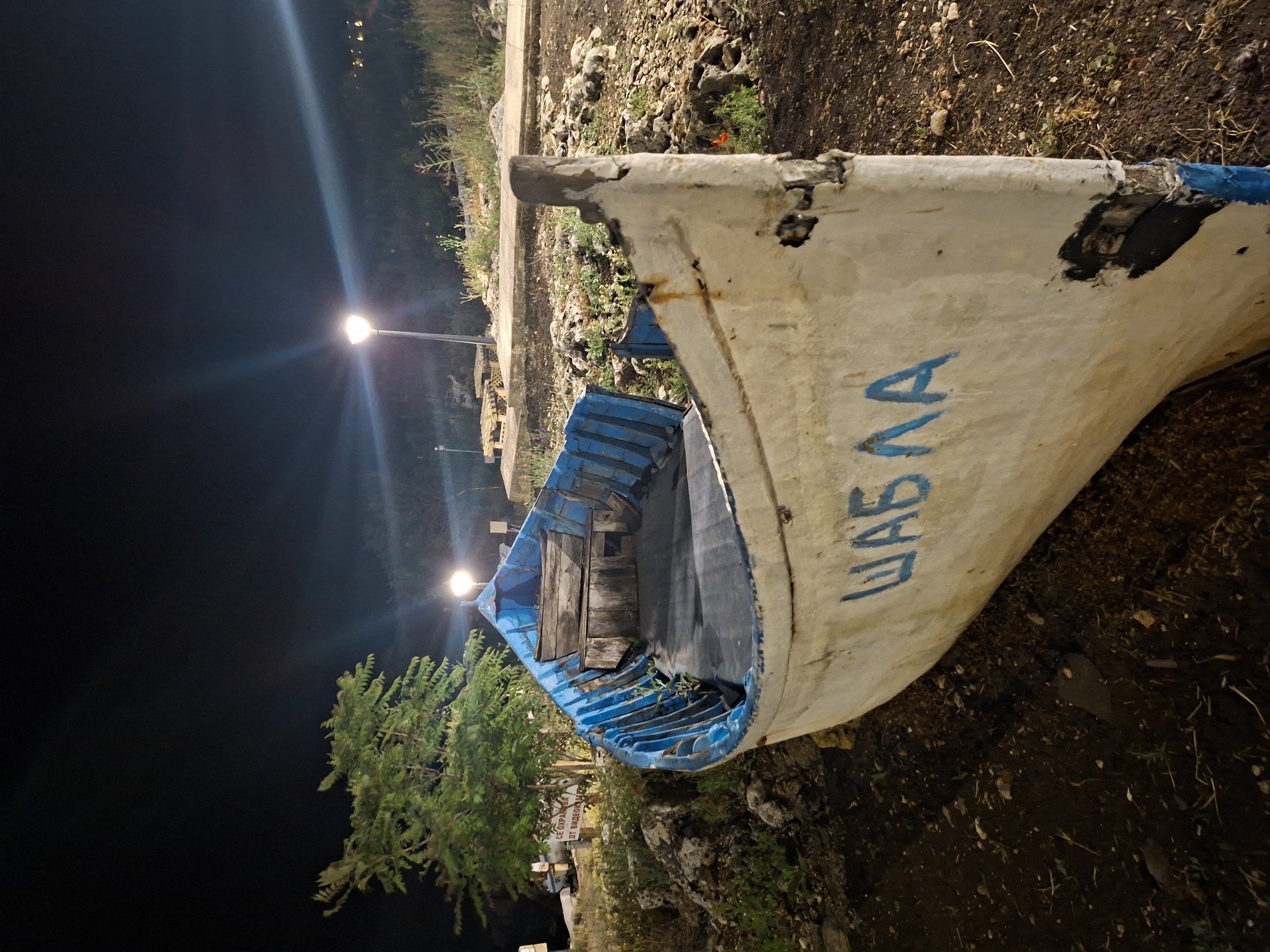 The image shows a weathered, white and blue fishing boat named "ШАБЛА" in the city of Tyulenovo, Bulgaria, at night. The boat is pulled ashore and rests on dark soil and rocks. Its blue interior is visible, with wooden benches and seats. The boat's hull shows signs of age and wear, with chipped paint and repaired sections. In the background, two streetlights illuminate the scene, casting long beams of light into the dark sky. Lush green vegetation, including a tree on the left, surrounds the boat and stretches up a gentle slope. The ground is covered with dirt, stones, and sparse grass.