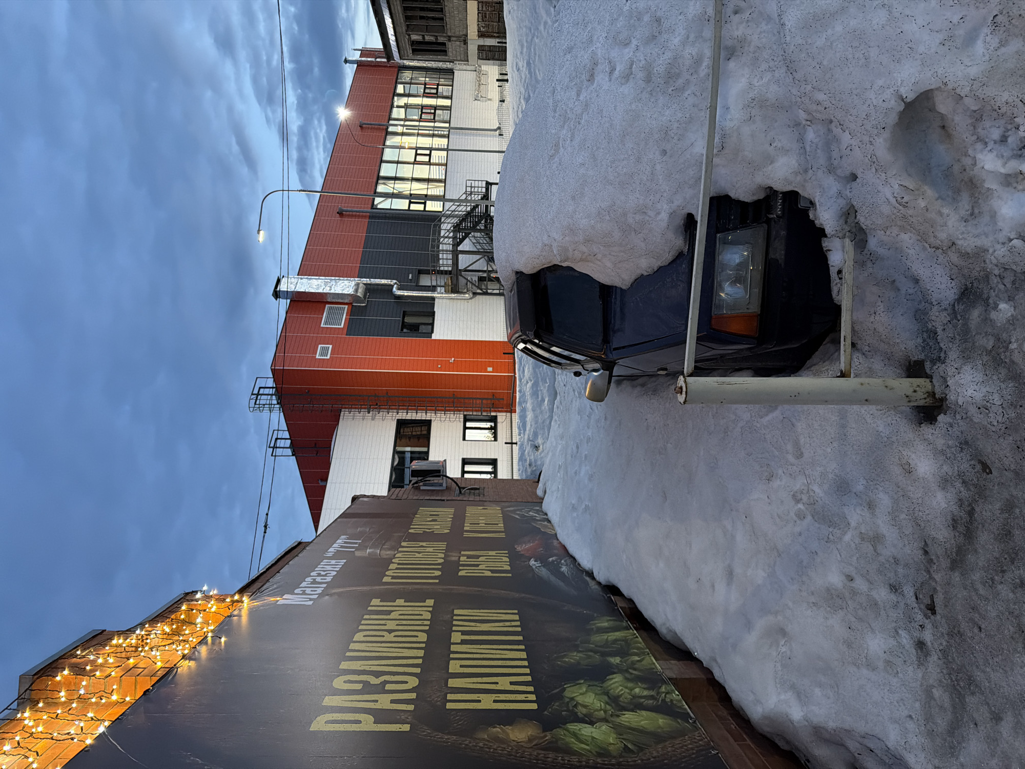 The image depicts a scene in Kodinsk, Russia. A dark-colored car is partially buried in a snowdrift. A billboard advertising "Razlivnye Napitki" (draft drinks), "Gotovaya Zakuska" (ready snacks), and "Ryba Kopchenaya" (smoked fish) with a picture of hop cones is partially visible in the foreground. In the background, there's a building with sections painted in red, grey, and white. The sky is overcast. A streetlight is visible near the building.