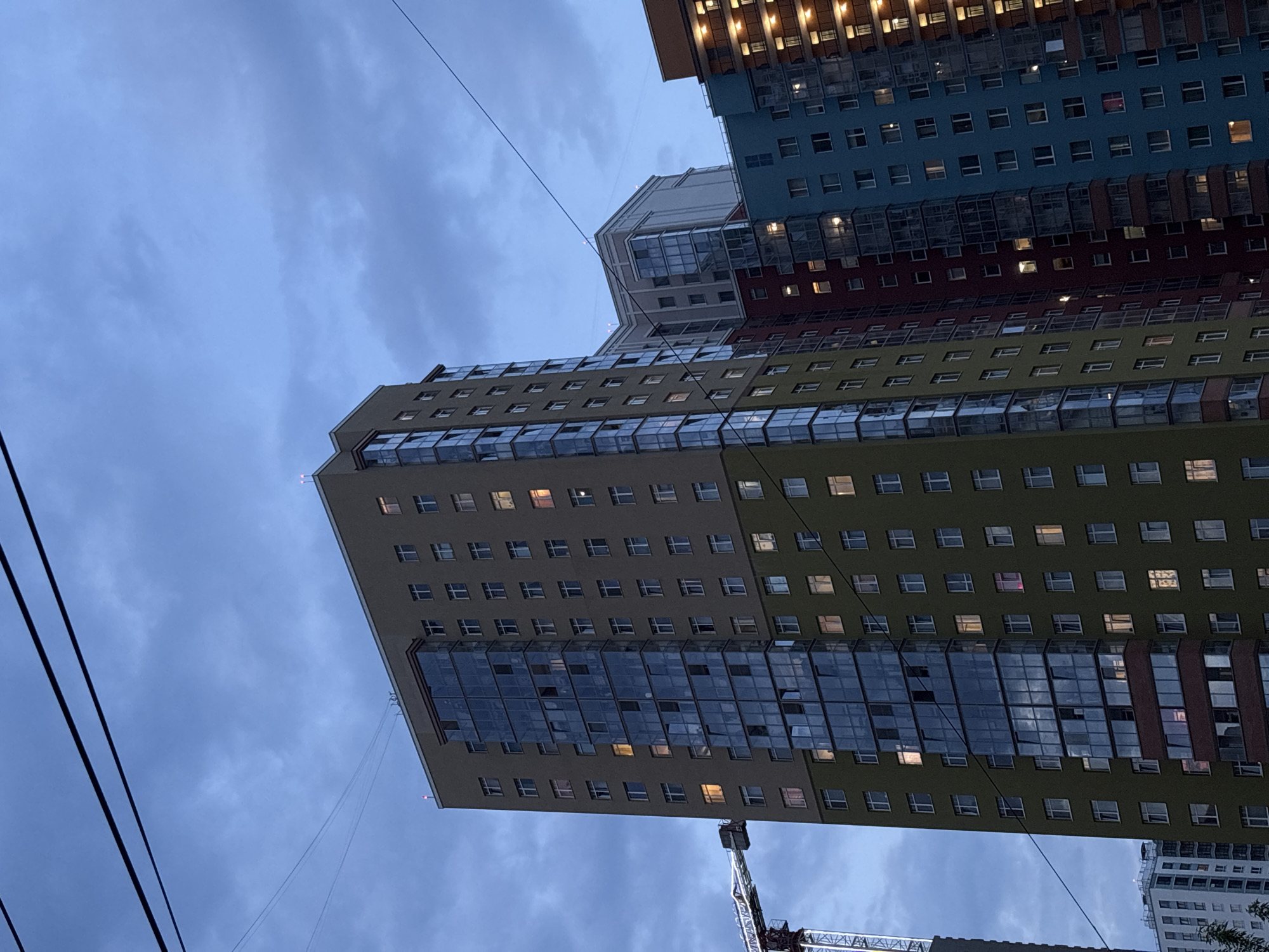 The image is an upward-looking shot of a group of modern, multi-story apartment buildings in Perm, Russia. The buildings are distinguished by their colorful facades, including blue, red, yellow, and green sections. Many windows are lit, suggesting that the apartments are occupied. Overhead, power lines stretch across the frame against a cloudy sky. A construction crane is visible in the lower portion of the image.
