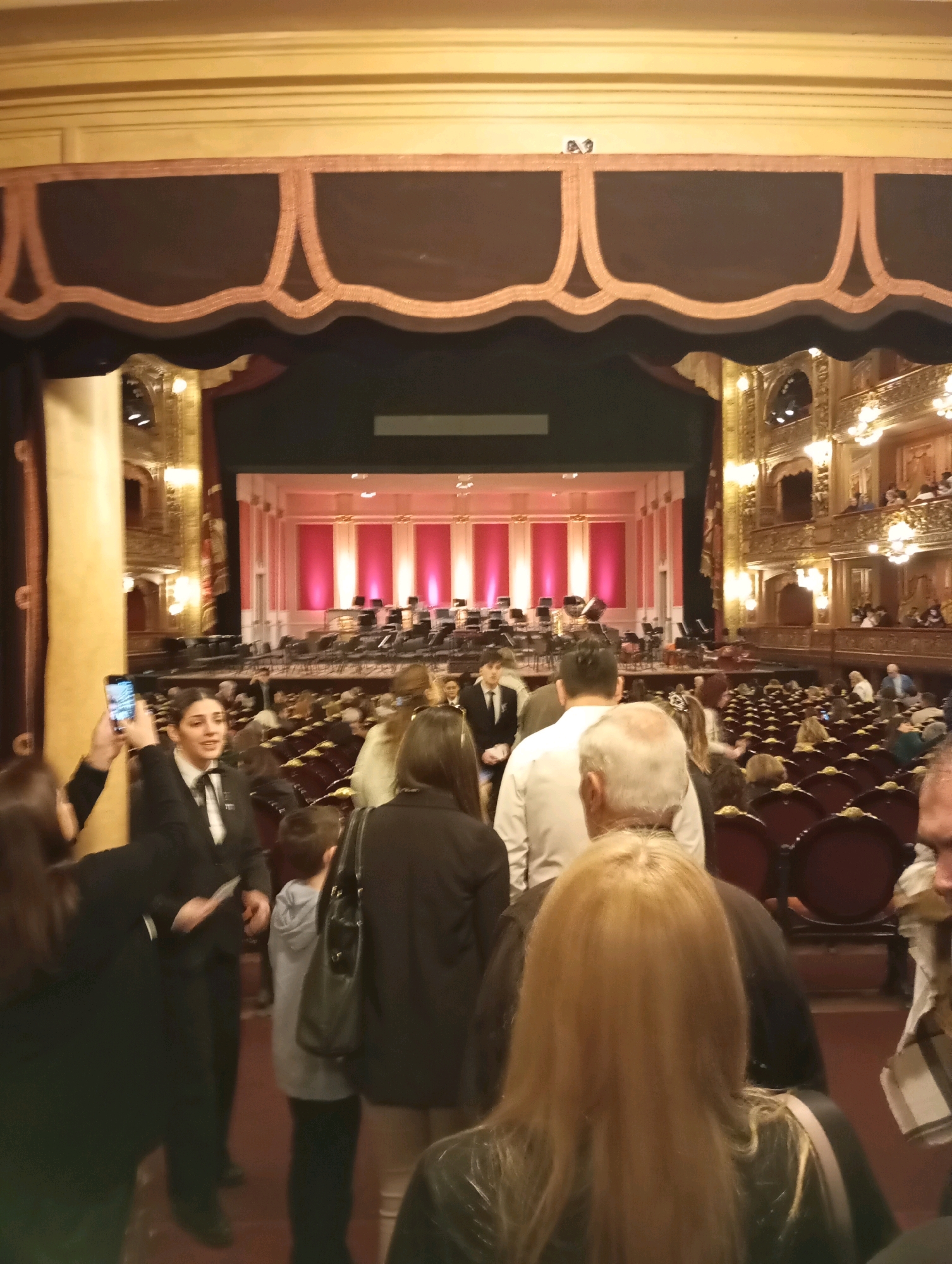 This image depicts the interior of a grand theater, likely in Buenos Aires, Argentina, given the context of an opera or symphony hall. The perspective is from within the audience seating area, looking towards the stage. The ornate architecture of the theater is evident, with gilded balconies, red velvet seating, and a richly decorated proscenium arch. The stage itself is set with rows of chairs for an orchestra, and pink-hued lighting illuminates the backdrop.
In the foreground and midground, a crowd of people are gathered, appearing to be patrons or staff. Some individuals are looking at their phones, while others are engaged in conversation or looking towards the stage. A woman in a black suit and white shirt, possibly an usher, is visible on the left, holding a phone up to take a picture. A young boy in a grey hoodie is standing near the center, and a group of people with blonde hair are prominent in the mid-right. The overall atmosphere suggests an event is about to begin or has just concluded. The lighting is warm and theatrical, contributing to the elegant ambiance of the venue.