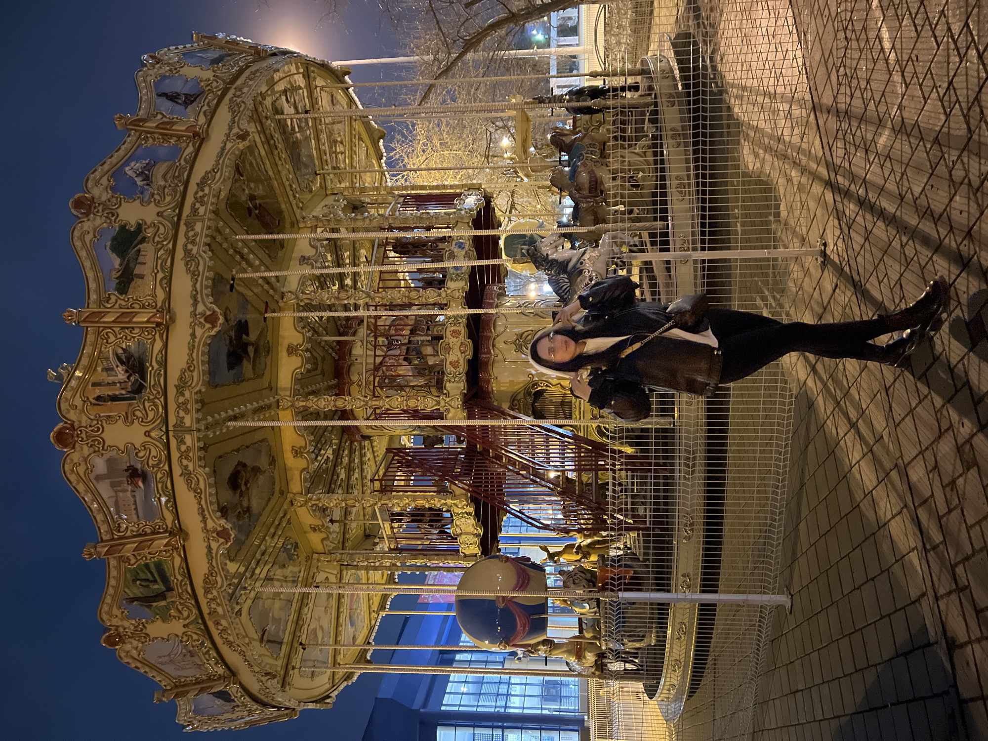 An eye-level outdoor shot shows a woman standing behind a white metal fence in front of an illuminated carousel at night. The carousel, ornately decorated in gold and cream colors, features depictions of various scenes. The woman is wearing dark clothing and a light-colored bag. The background includes a dark blue night sky, city lights, and a brick walkway. The scene is in Tyumen, Russia.