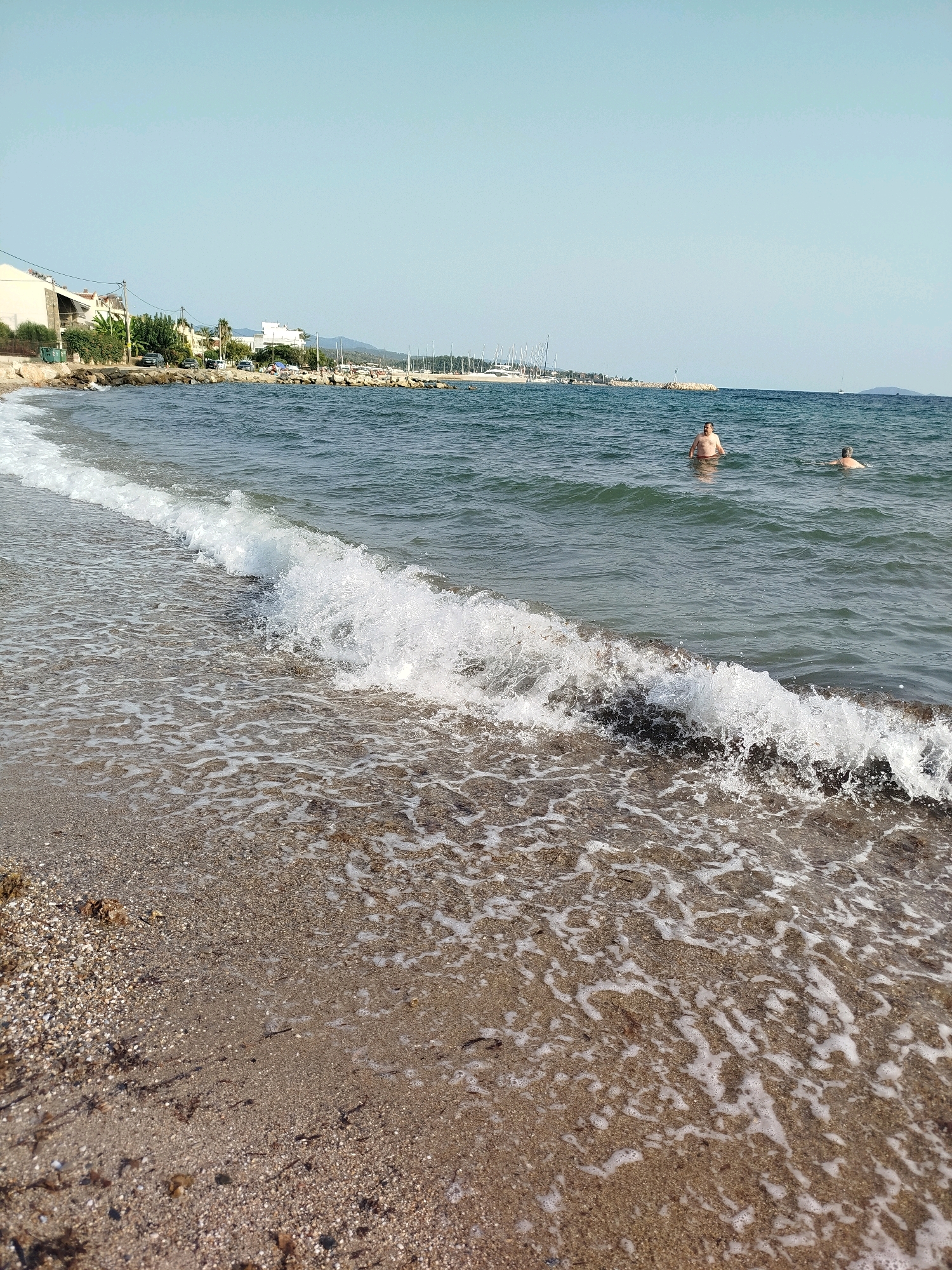 The image depicts a coastal scene in Sithonia Municipal Unit, Greece, on a clear, sunny day. The foreground shows a beach with fine-grained sand and small pebbles, with the gentle waves of the sea lapping at the shore, creating white foam and ripples. In the middle ground, two men are visible in the water, appearing to be bathing or swimming. The water is a deep blue-green, with some choppiness. In the background, a line of buildings, trees, and a road runs along the coastline. Beyond the buildings, a harbor filled with sailboats and yachts is visible, indicating a populated area. The sky is a pale, cloudless blue. The overall impression is a peaceful, recreational day at the seaside.