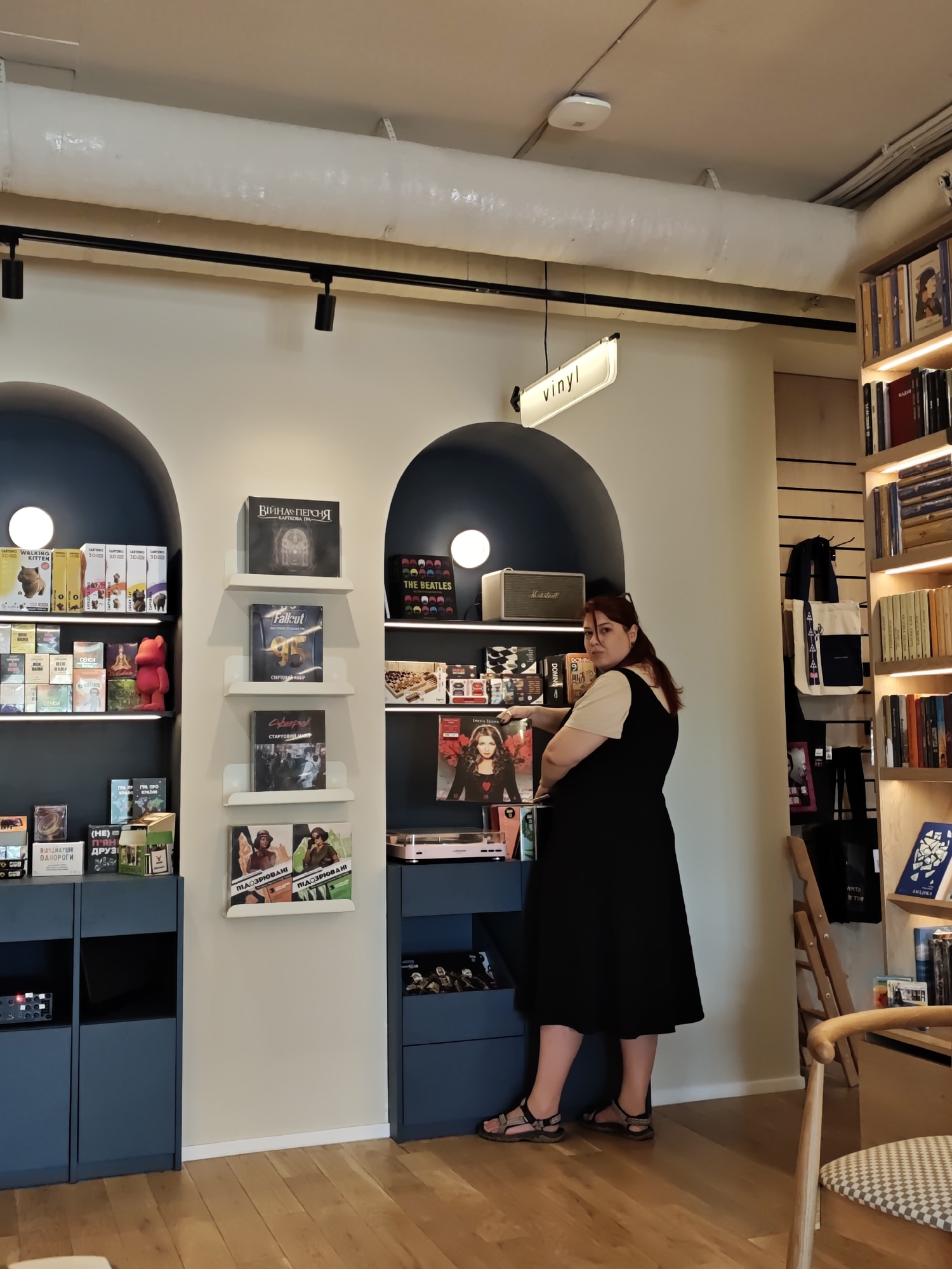 The image depicts the interior of a vinyl record store in Kyiv, Ukraine. A woman with dark red hair, wearing a black dress over a beige top and sandals, stands in the middle of the frame. She is looking towards the right side of the image and is holding a vinyl record. The store features shelves filled with vinyl records, books, and other items. In the background, there are arched shelves displaying various records and a "vinyl" sign is visible on the ceiling. The floor is made of wood. The lighting suggests an indoor retail environment.