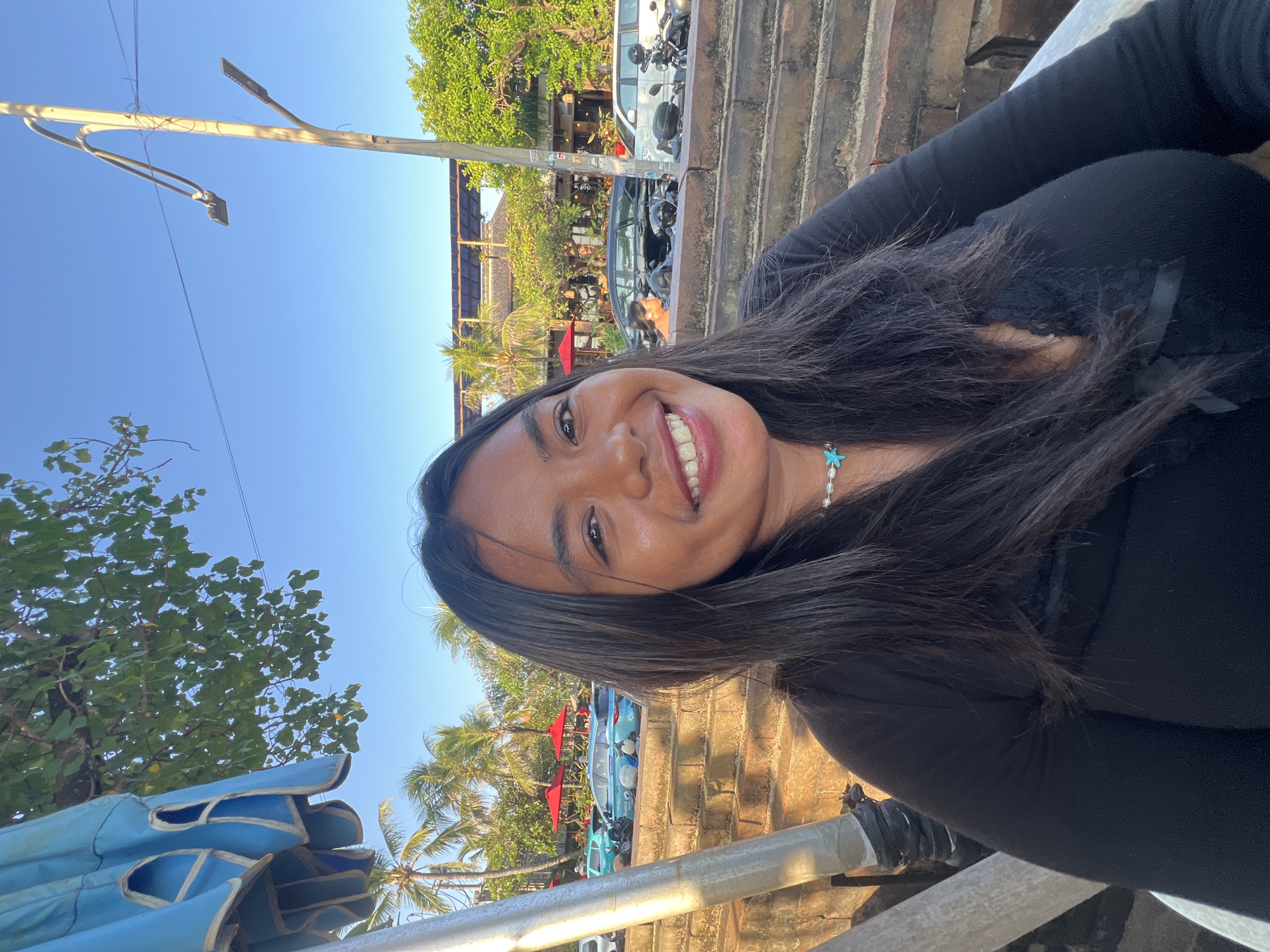 In Kuta, Indonesia, a young woman with long, dark hair smiles at the camera in a close-up portrait. She is wearing a black long-sleeved top with lace detailing and a pearl necklace with a turquoise starfish pendant. The background reveals a street scene with palm trees, buildings, parked cars, and red umbrellas. The lighting suggests it is late afternoon or early evening.