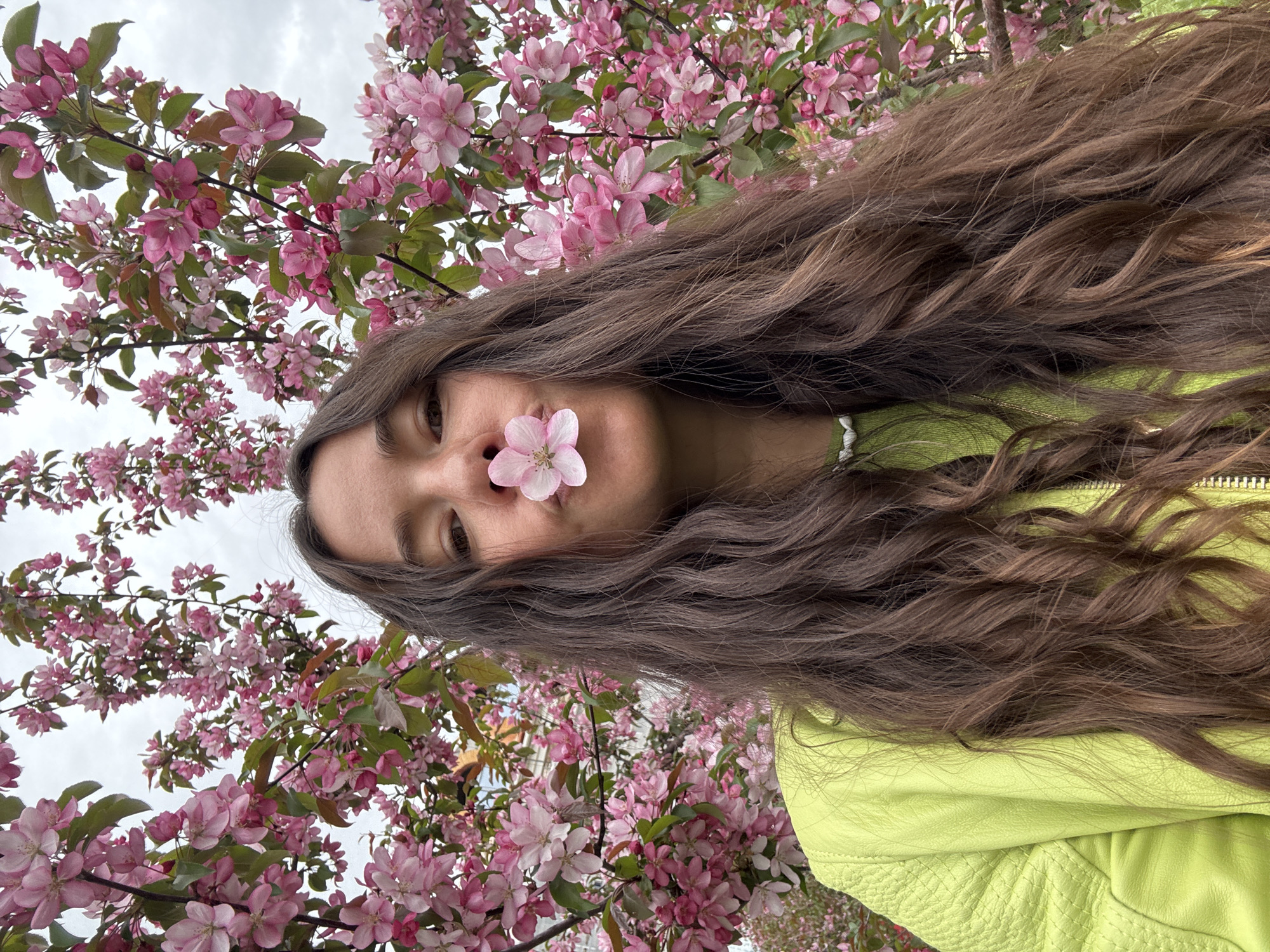 A young woman with long, wavy brown hair poses with a pink flower held to her nose. She wears a bright lime-green jacket. The background features a profusion of pink blossoms and green leaves, suggesting a flowering tree. The sky is overcast. While the prompt mentions Kazan, Russia, there is no visual evidence in the image to confirm the location.