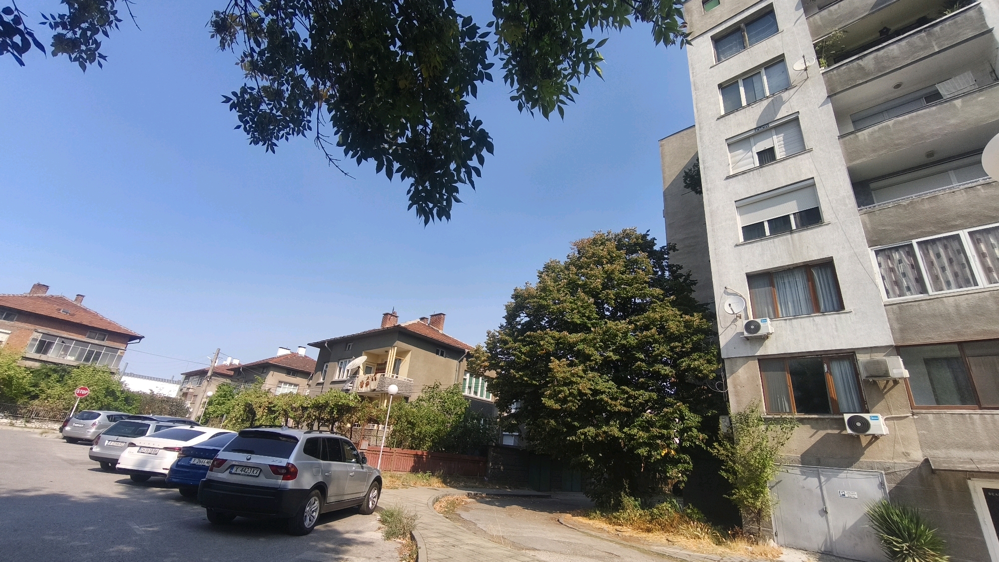 The image depicts a street scene in Dimitrovgrad, Bulgaria, on a clear, sunny day. In the foreground, a paved sidewalk runs alongside a street with parked cars. Several vehicles are visible, including a silver BMW X3 in the right foreground. Behind the parked cars, residential buildings of varying architectural styles are present, featuring tiled roofs and balconies. One building on the right is a tall, multi-story apartment complex with numerous windows and air conditioning units. On the left, a smaller, older building with a reddish-brown tiled roof is visible. Lush green trees, some with autumn foliage, are interspersed throughout the scene, with branches of a tree hanging from the top of the frame. The sky is a clear, bright blue. There are no people visible in the image. The overall impression is one of a quiet urban residential area.