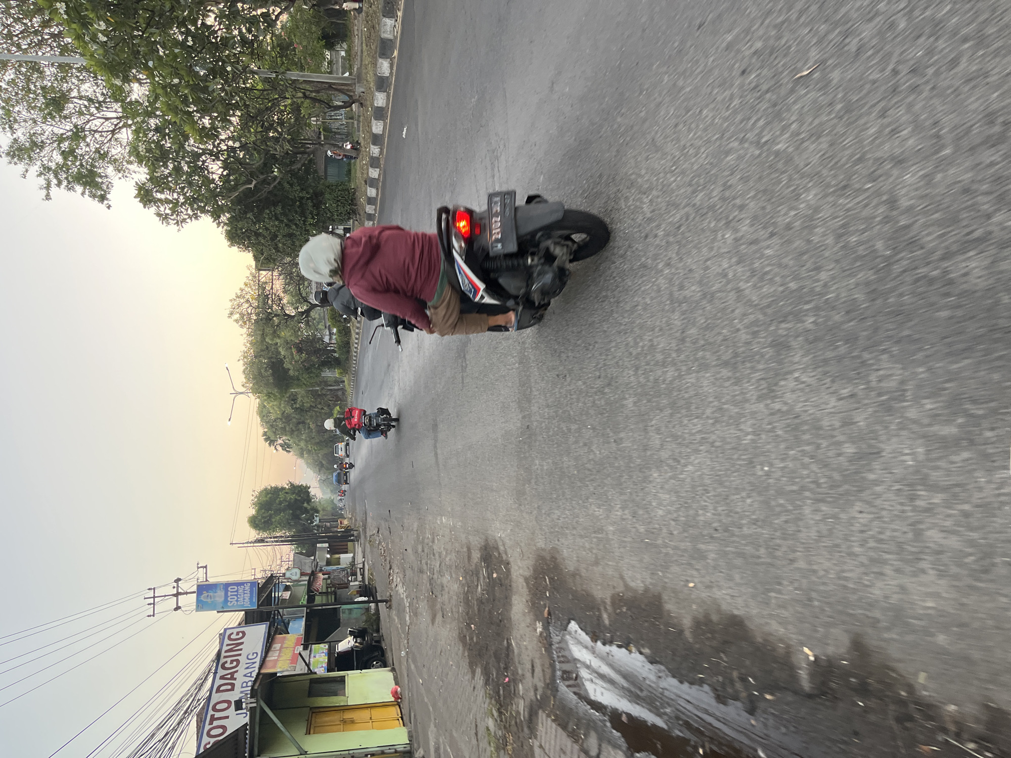 The image depicts a street scene in Ngaban, Indonesia, likely during the early morning or late afternoon given the soft, warm light and hazy sky. The foreground is dominated by a paved road with wet patches, suggesting recent rain or morning dew. A person wearing a helmet and a maroon hoodie rides a scooter in the mid-ground, facing away from the camera. They are also wearing brown pants and flip-flops. The scooter has a red taillight illuminated and a license plate that reads "W 2702 SJY".
To the left of the road, there is a row of small shops or buildings. One building prominently displays a sign in Indonesian that reads "SOTO DAGING JOMBANG" (Beef Soto Jombang), indicating a food establishment. Another smaller sign above it shows a blue image of a bottle with a white label and text. A utility pole with numerous wires is also visible near the buildings.
Further down the road, several other vehicles, including cars and scooters, are moving in the same direction. Trees line the right side of the road, with some appearing to be flowering. The overall impression is one of a typical urban or semi-urban environment in Indonesia during a low-light period of the day.