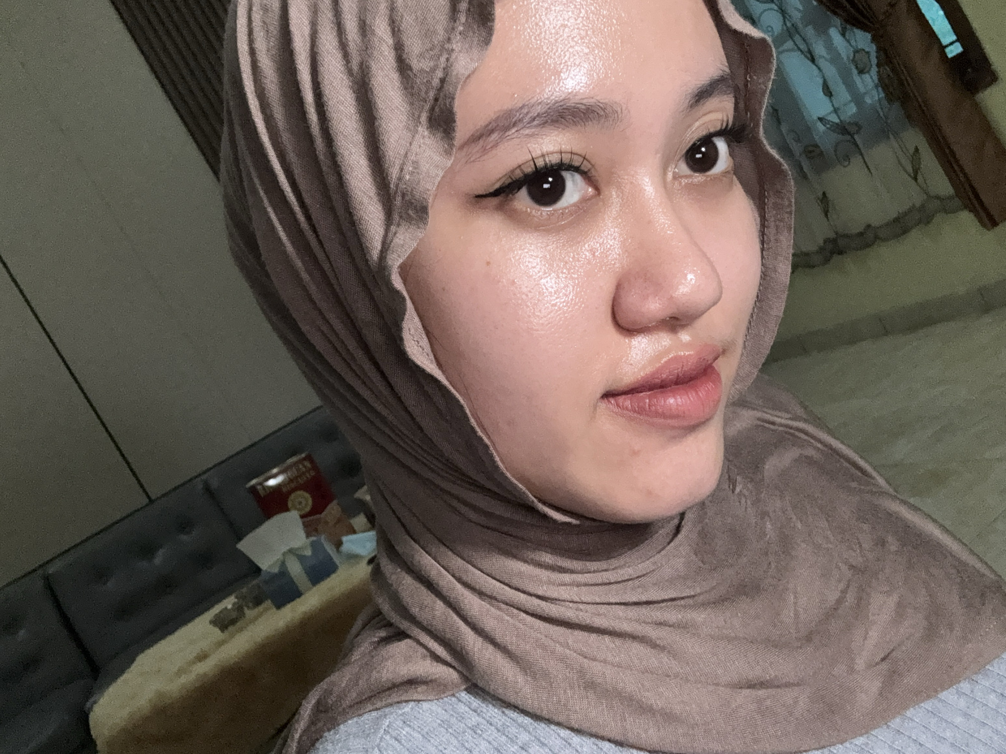 A close-up photograph shows a person wearing a light brown hijab, with their face turned slightly to the right. The person has fair skin, dark eyes with eyeliner, and full lips. The fabric of the hijab is draped around the person's head and shoulders. In the background, there is a gray couch with a dark brown wall, and a table with various objects on it, including a red can or box. The photo appears to have been taken indoors. There is no information provided in the prompt that suggests a location in the city of Gadingrejo, Indonesia.