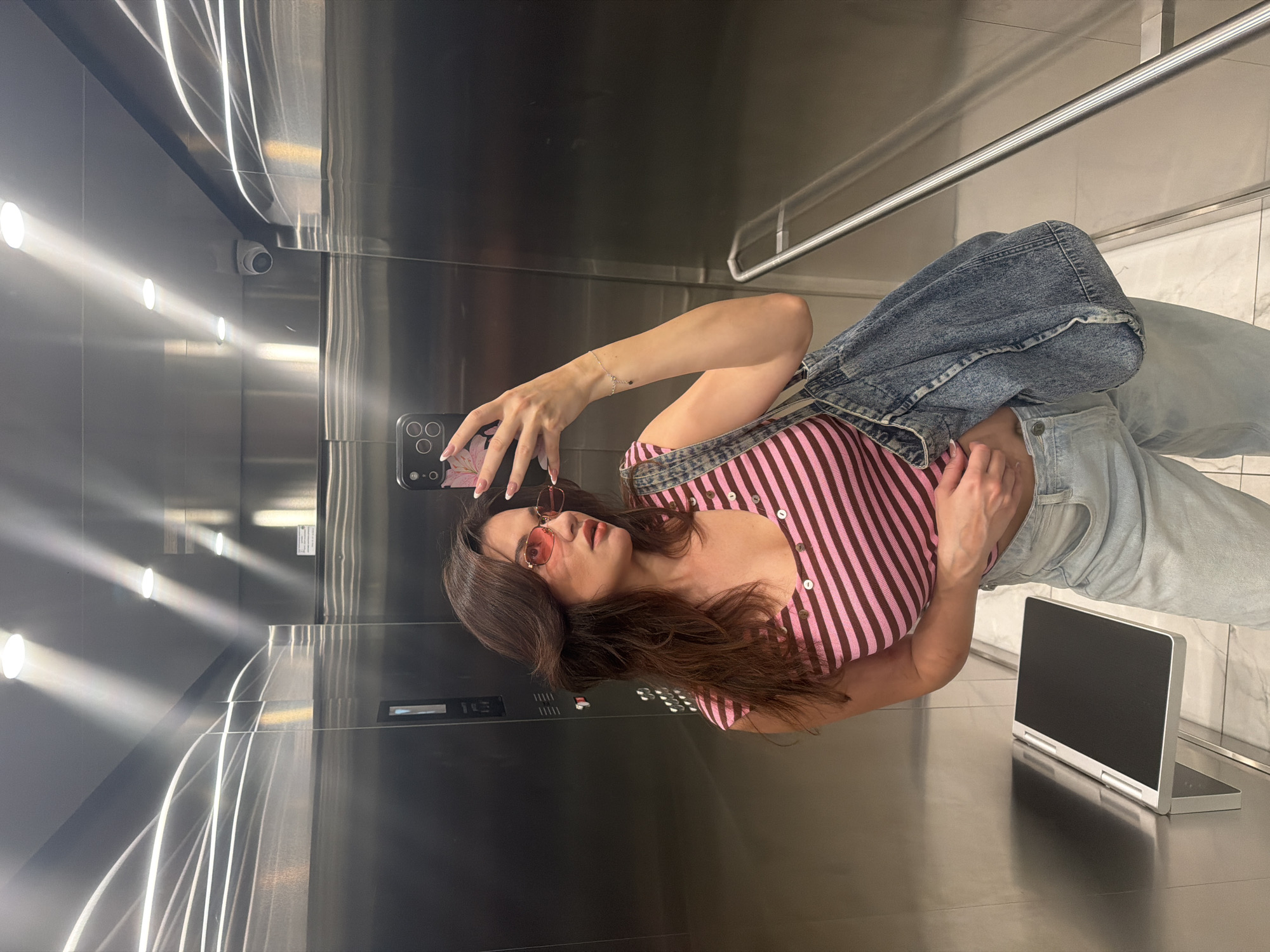 A woman is taking a selfie in an elevator. She is wearing a pink and brown striped button-up shirt, blue jeans, and a denim jacket. She has brown hair and is wearing red sunglasses. The elevator has stainless steel walls and a white marble floor. A laptop is standing on the elevator floor. The location appears to be in Belgrade, Serbia, although this cannot be confirmed solely from the image.