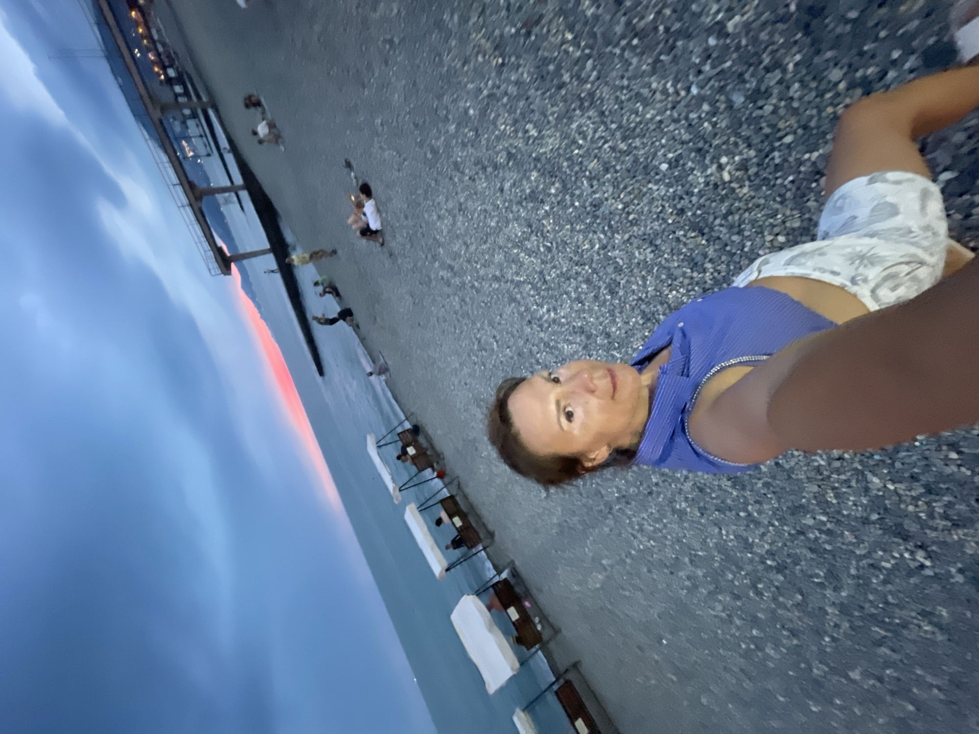 In the city of Khostinskiy, Russia, at twilight, a woman is captured in a selfie on a pebbled beach. She wears a blue, sleeveless top and white patterned shorts. The sea stretches out behind her, with a sunset painting a pink and orange hue across the horizon. A pier with lights is visible in the distance, and several people are scattered on the beach, some sitting on swings with canopies, while others recline on the shore. The sky is a deep blue, with wispy clouds.