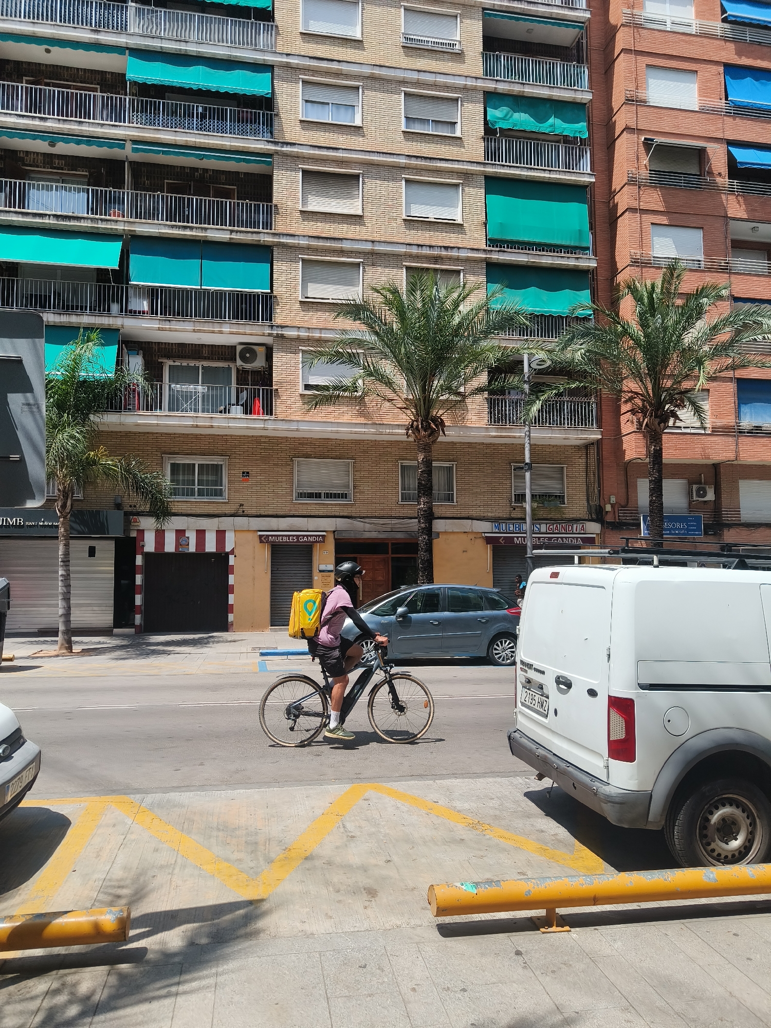 A person on a bicycle wearing a helmet and a pink shirt with a yellow backpack rides along a street in Gandia, Spain. The cyclist is positioned in the middle of the frame, with the bicycle's wheels on the asphalt road. To the right, a white van is parked, its rear visible. The background features a multi-story brick building with balconies and teal awnings, and palm trees line the sidewalk. The street is marked with yellow painted lines on the pavement. The scene appears to be a sunny day.