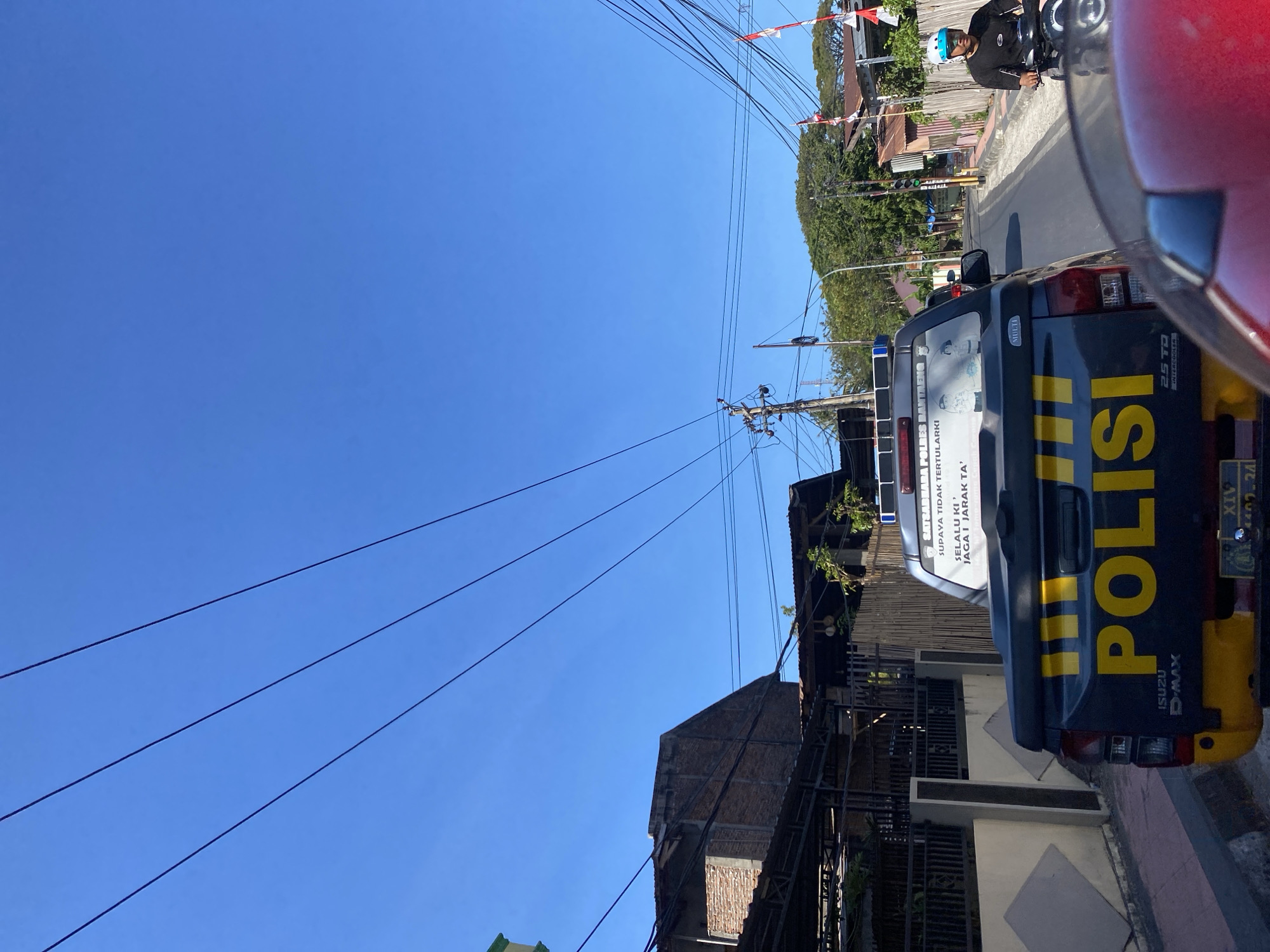 The image shows a rear view of a black police utility truck with yellow "POLISI" lettering on the tailgate, parked on a street in Bantaeng, Indonesia. The truck has a blue light bar on the roof and a license plate that reads "B 1102 ZAJ". A sticker on the back of the truck reads "SATGAS PANTAS BANTEENG" and "UPAYA TIDAK TERTULARI" followed by "SELALU KI JAGA I JARAK TA'". To the right of the truck, a man wearing a black shirt and a blue helmet rides a motorcycle. The street is paved and lined with buildings and trees. Overhead, several power lines are visible against a clear blue sky.