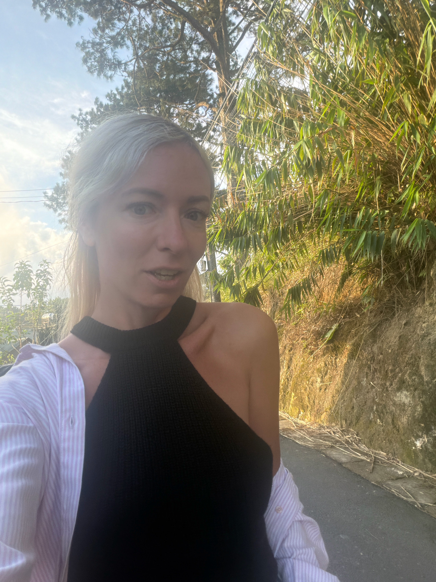 A medium shot photo features a fair-skinned woman with blonde hair pulled back in a ponytail, wearing a black halter top and an unbuttoned pink and white striped shirt. She is looking towards the camera with a slight smile and her mouth open as if speaking. The background shows a paved road next to a rocky embankment covered in dry grass and bamboo plants. A large tree with sparse branches is visible in the upper left corner. The scene appears to be in a rural or mountainous area, possibly on the outskirts of Xuân Trường, Đà Lạt, Vietnam, given the vegetation and terrain. The lighting suggests it is daytime, with the sun casting a warm glow on the scene.