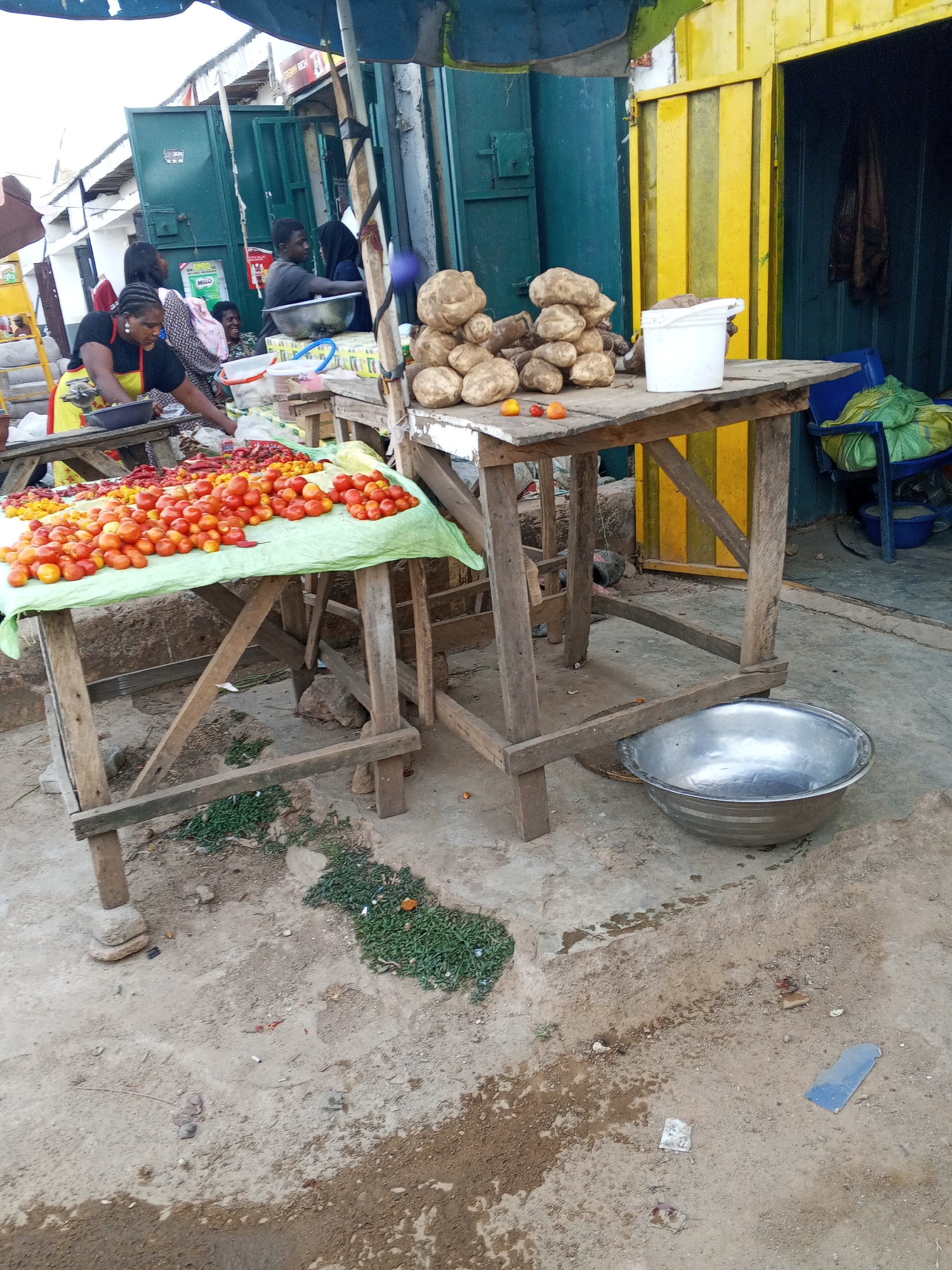 This image captures a bustling outdoor market scene in Jos, Nigeria, showing various market activities and produce displays.
In the foreground, two rustic wooden tables serve as display stands. The table on the left is covered with a light green cloth and laden with a large assortment of fresh tomatoes, predominantly red and orange, along with some red chili peppers. A woman, wearing a black top and a yellow apron, is actively sorting or arranging the produce on this table. Other people are visible in the background, including a man in a black shirt and a woman with a head covering, seemingly engaged in transactions or market work near a stall.
The table on the right holds a large pile of what appear to be yams or similar root vegetables, along with a white plastic bucket. A large, shiny silver bowl rests on the dirt ground beneath this table. Behind the tables, market stalls or buildings are visible. One structure has dark green doors or shutters, while another features a distinctive yellow and black striped door. A blue plastic chair with a green sack on it is placed near the yellow door. A blue canopy partially covers the area from above.
The ground is unpaved, consisting of dirt and sand with patches of small green plants and some scattered debris. The scene suggests a typical daytime market, busy with vendors and customers, highlighting the daily commerce in this region. A small green sign with the word "MILO" is visible in the background.