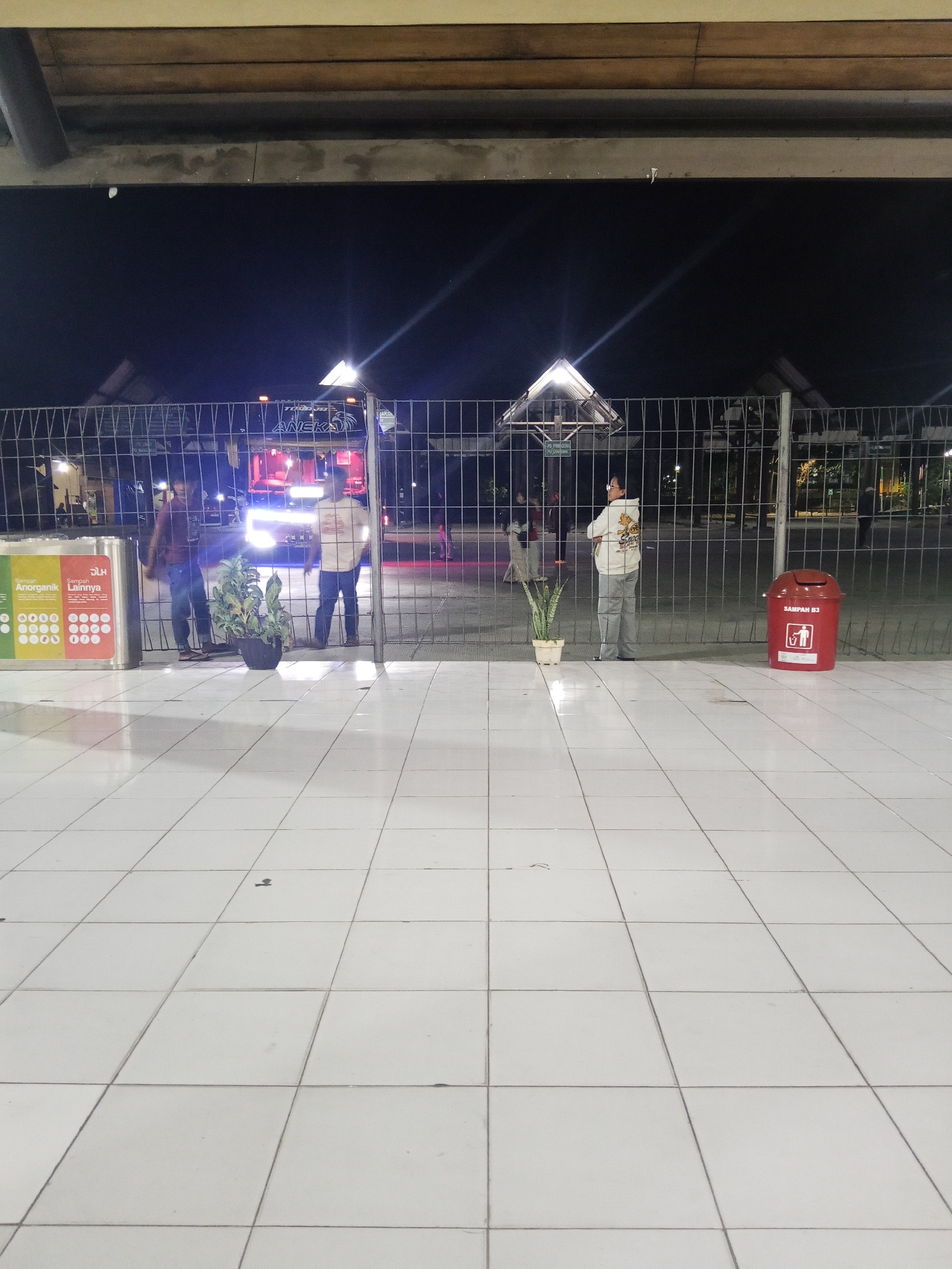 The image shows a street scene at night, likely in Makassar, Indonesia, near a bus station or transportation hub. A white tiled floor extends into the foreground. A metal fence separates the foreground from the background, where a bus with illuminated headlights and signs is visible. Two men are standing behind the fence. To the left, a multi-compartment trash receptacle is labeled with "Sampah Anorganik" and "Sampah Lainnya". To the right, a red trash bin labeled "SAMPAH B3" is present. A potted plant is placed near the fence, between the two men and the trash receptacle. The environment appears to be outdoors, with artificial lighting illuminating the scene.