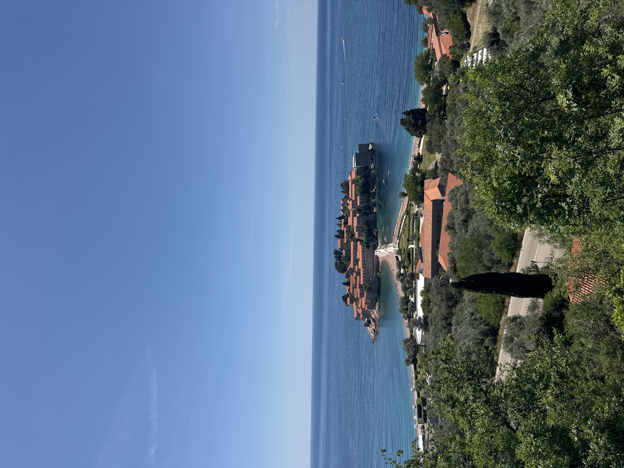 The image depicts an aerial view of Sveti Stefan, Montenegro, from a high vantage point. Sveti Stefan is a small islet with terracotta-roofed buildings clustered together. The island is connected to the mainland by a narrow isthmus with a sandy beach visible on either side. The Adriatic Sea surrounds the islet, with several small boats visible in the water. Lush green vegetation covers the mainland, including trees and bushes. A road runs along the coast on the mainland. The sky is clear and blue.