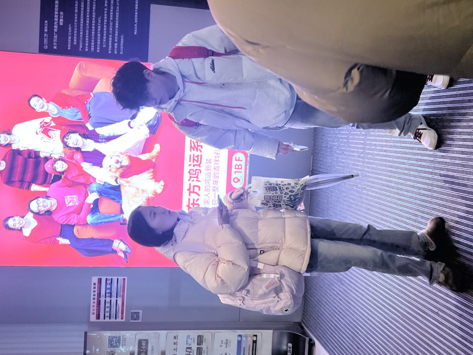 The image depicts an indoor scene within a commercial building, likely a mall, in Jiangjin, China. Two young adults, an East Asian female and an East Asian male, stand facing each other on a dark grey ribbed floor matting, suggesting an interaction.
The female, positioned on the left, wears a light pink puffer jacket, light blue jeans, and dark, bulky shoes. She carries a light pink backpack on her left shoulder. Her right hand holds a smartphone, while her left hand holds a clear plastic bag containing a clear umbrella. She has short, dark hair and is looking towards the male subject.
The male, positioned on the right, wears a light grey hoodie layered under a white puffer vest, light blue jeans, and white sneakers with red accents. He has a red backpack on his shoulders and holds a smartphone in his right hand. His dark hair is styled in a shaggy manner. He is looking towards the female subject.
In the background, a large, brightly illuminated digital display features an advertisement with a group of diverse individuals and a golden retriever dog, all dressed in red tones and sitting on red furniture. Chinese characters are visible on the display, including "东方鸿运系" and "1BF," indicating a commercial context and floor level. To the far left, a white information stand displaying QR codes and additional Chinese text is partially visible. A third person, mostly obscured, is partially visible in the far right foreground, wearing a dark puffer jacket and jeans. The scene is illuminated by artificial lighting.