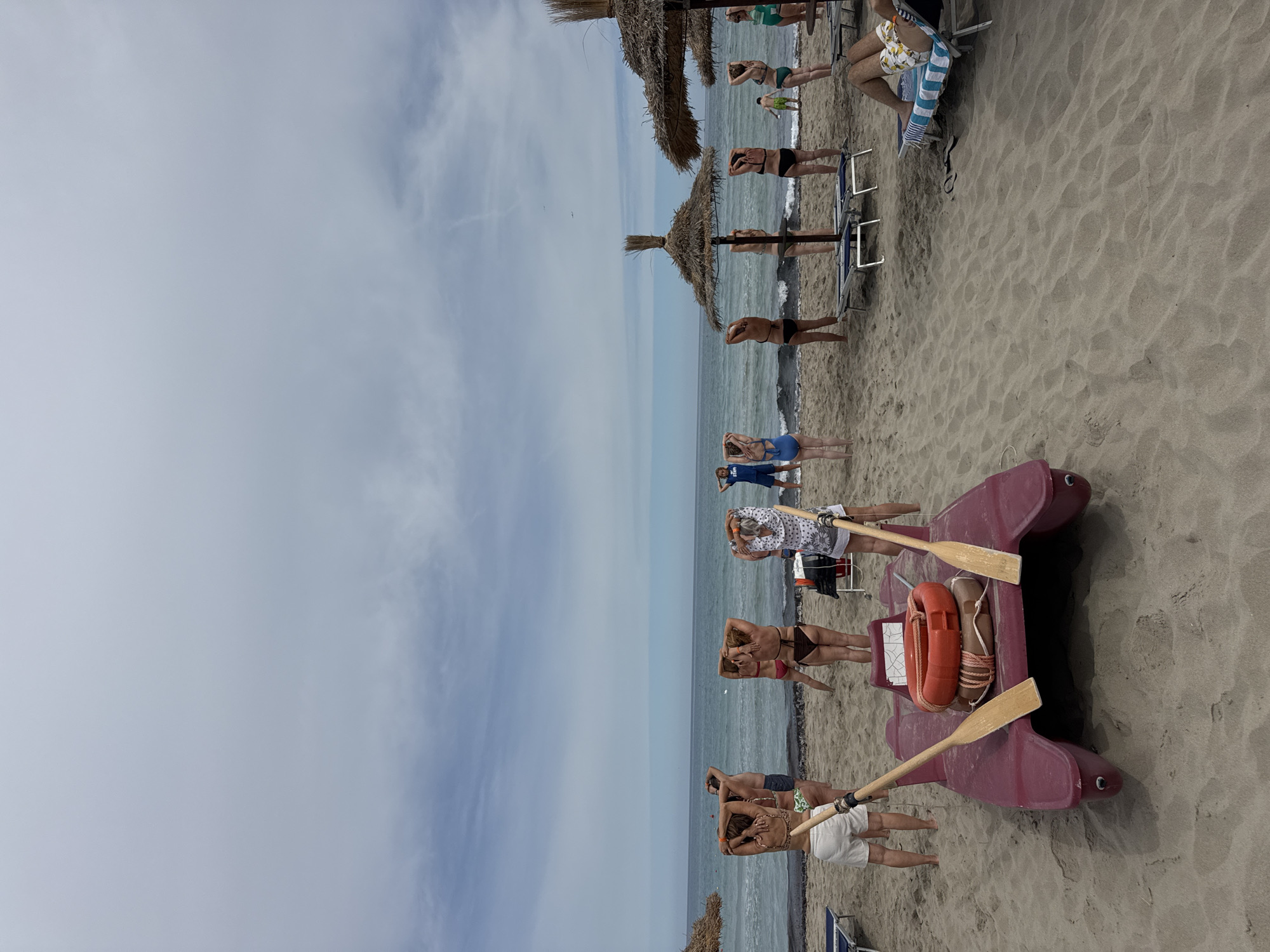 In Isola delle Femmine, Italy, on a sandy beach under a cloudy sky, a group of people are performing exercises near the water. A red, two-person pedal boat with two wooden oars lies on the sand in the foreground. The boat is equipped with an orange life preserver and a rope. Several individuals, dressed in swimwear, are positioned facing the sea with their arms behind their heads, engaged in stretching or a similar activity. Beach umbrellas and lounge chairs are visible in the background. A person in a blue shirt with "STAFF" printed on it stands further down the beach. The sea is calm with small waves.
