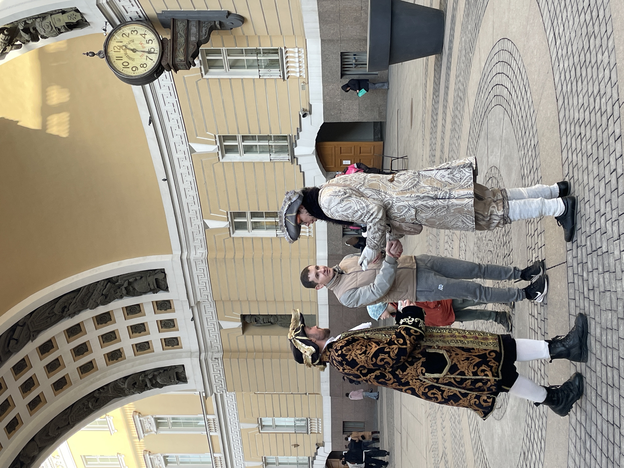 The image depicts an outdoor scene in Saint Petersburg, Russia, specifically in Palace Square, with the General Staff Building and its archway serving as the prominent background structure.
In the foreground and midground, three adult males are interacting on a paved square. The man on the left wears historical period attire consisting of a black and gold brocade long coat, white stockings, and black boots. He has a dark hat with gold trim. The man in the center wears modern casual clothing: a beige padded vest over a grey long-sleeve shirt, grey sweatpants with a 'NIKE' logo, and black sneakers. He holds a white cup in his left hand. The man on the right also wears historical period attire, featuring a light-colored (possibly silver or cream) brocade long coat with a paisley pattern, light breeches, and a grey tricorn hat with dark fur trim. The man in modern attire (center) is shaking hands with the man in the light-colored period costume (right).
The background consists of the light-yellow, multi-story General Staff Building, characterized by numerous white-framed windows and decorative white architectural elements, including cornices and pilasters. A large arched passage, adorned with sculptural reliefs and coffered ceilings, is visible on the left. To the right, a large, ornate clock with Roman numerals, displaying approximately 3:45, is mounted on the building facade. Below the clock face, the numbers "765" are visible.
Other individuals are scattered in the background: a child in a red jacket behind the central male, a group of people to the far left, and a person in a dark hooded garment leaning against the building on the far right, looking down at a bright blue object. A large, dark-grey cylindrical planter is situated on the right side of the foreground. The ground is paved with light-colored pavers arranged in geometric and circular patterns.
