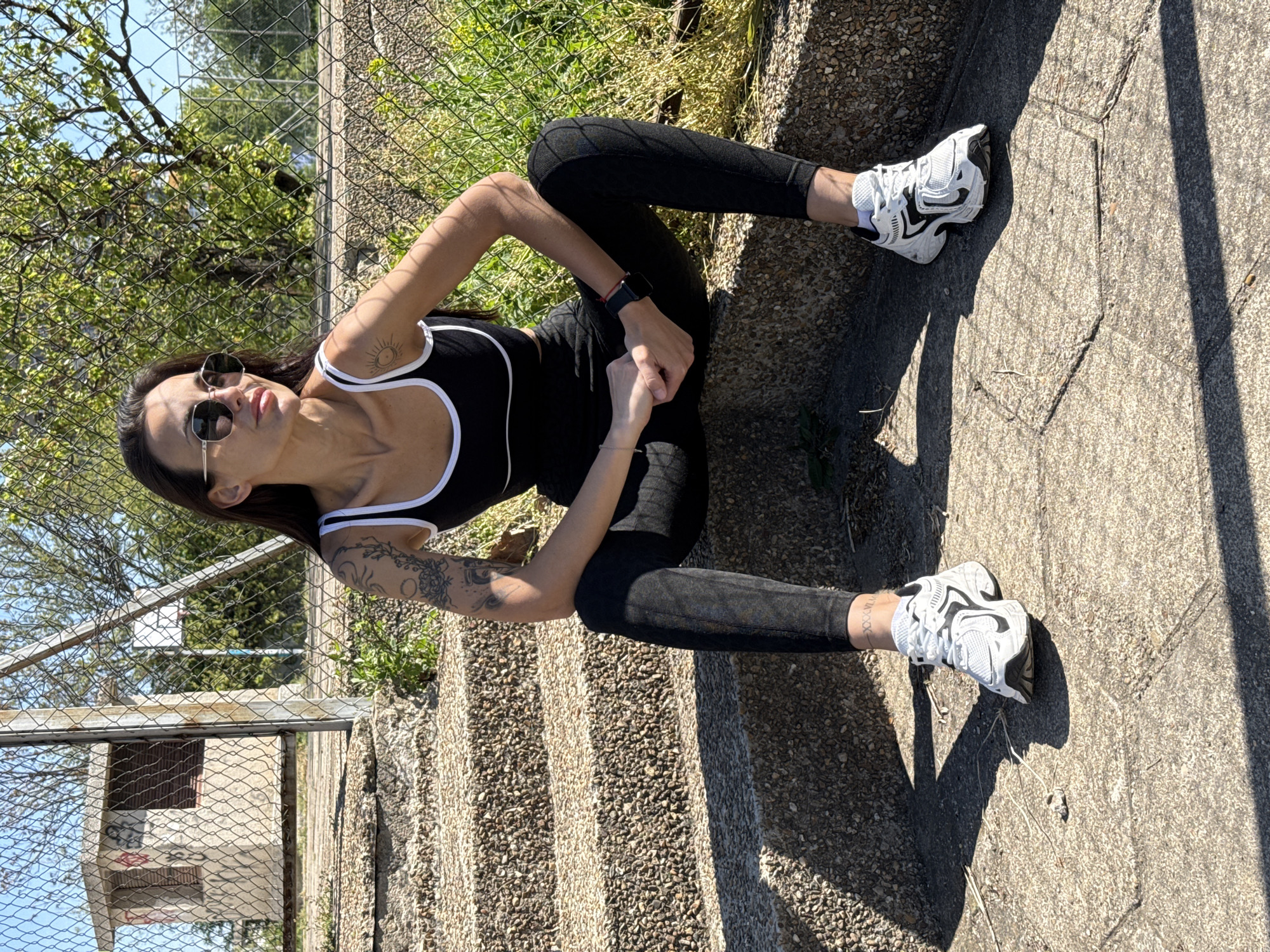 A woman is posing outside on a partly sunny day. She is wearing sunglasses, a black top with white trim, black leggings, and white and black athletic shoes. She has tattoos on her left arm, right wrist, and left collarbone area. She is sitting on a set of steps next to a chain-link fence, and behind the fence is a small building. The location is inferred to be Pleven, Bulgaria.