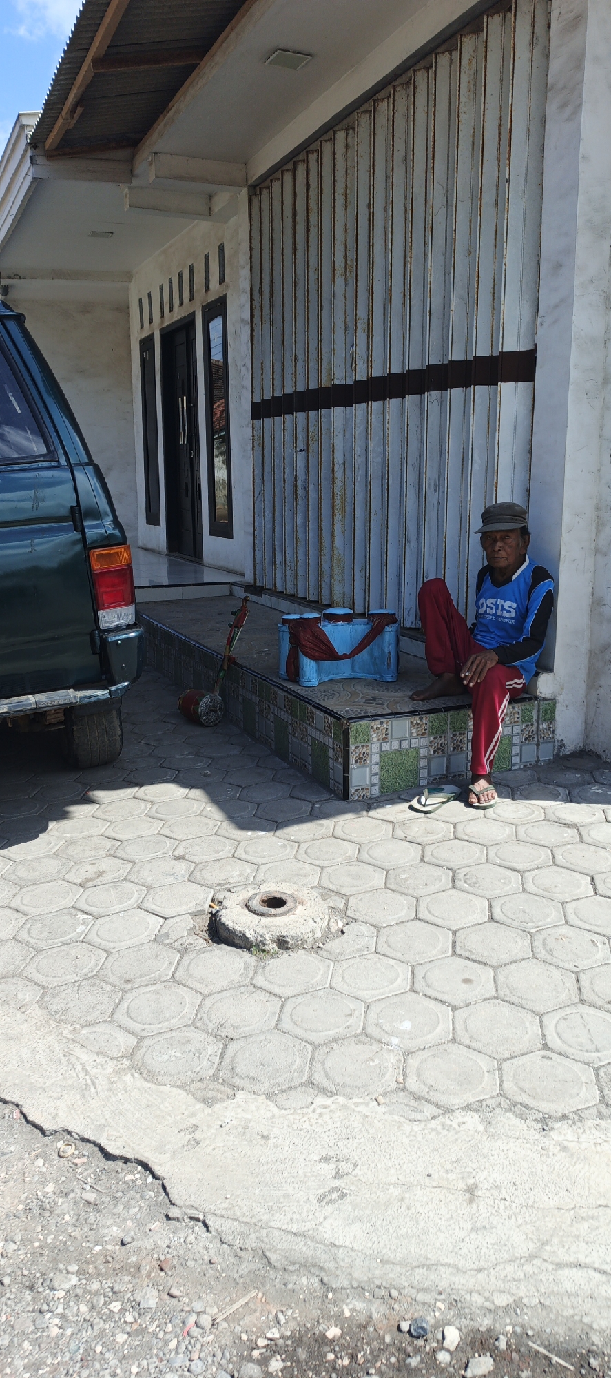 An elderly man sits on a tiled step outside a building with a large, corrugated metal roll-up door. He wears a grey cap, a blue shirt with "OSIS" printed on it, and red pants. A pair of white and green flip-flops are near his feet. Beside him is a blue object with a red cloth draped over it, and a toy that appears to be a drum or percussion instrument with a wheeled base. In the foreground, hexagonal paving stones form the ground surface, and a small concrete structure with a circular opening is visible. A portion of a dark green vehicle is parked to the left of the frame. The setting appears to be an outdoor area in Puger, Indonesia, characterized by its sunny weather and paved surfaces.