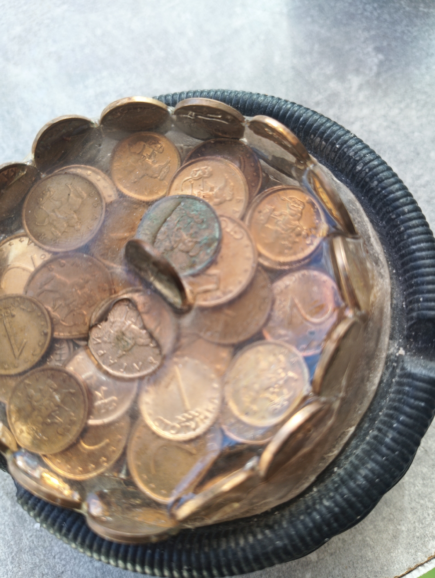 A close-up, high-angle shot shows a collection of coins embedded in clear resin within a black, ribbed bowl. The coins are mostly copper-toned, with some appearing tarnished or greenish. They are densely packed and overlap each other, creating a textured surface. The resin appears to be slightly cloudy in places. The bowl has a wide, black, ribbed rim and rests on a textured gray surface, possibly a concrete or stone countertop, which is partially visible in the background. The lighting suggests an outdoor or well-lit indoor setting. The image appears to have been taken in Karnobat, Bulgaria, although there are no explicit visual cues in the image to confirm this location.