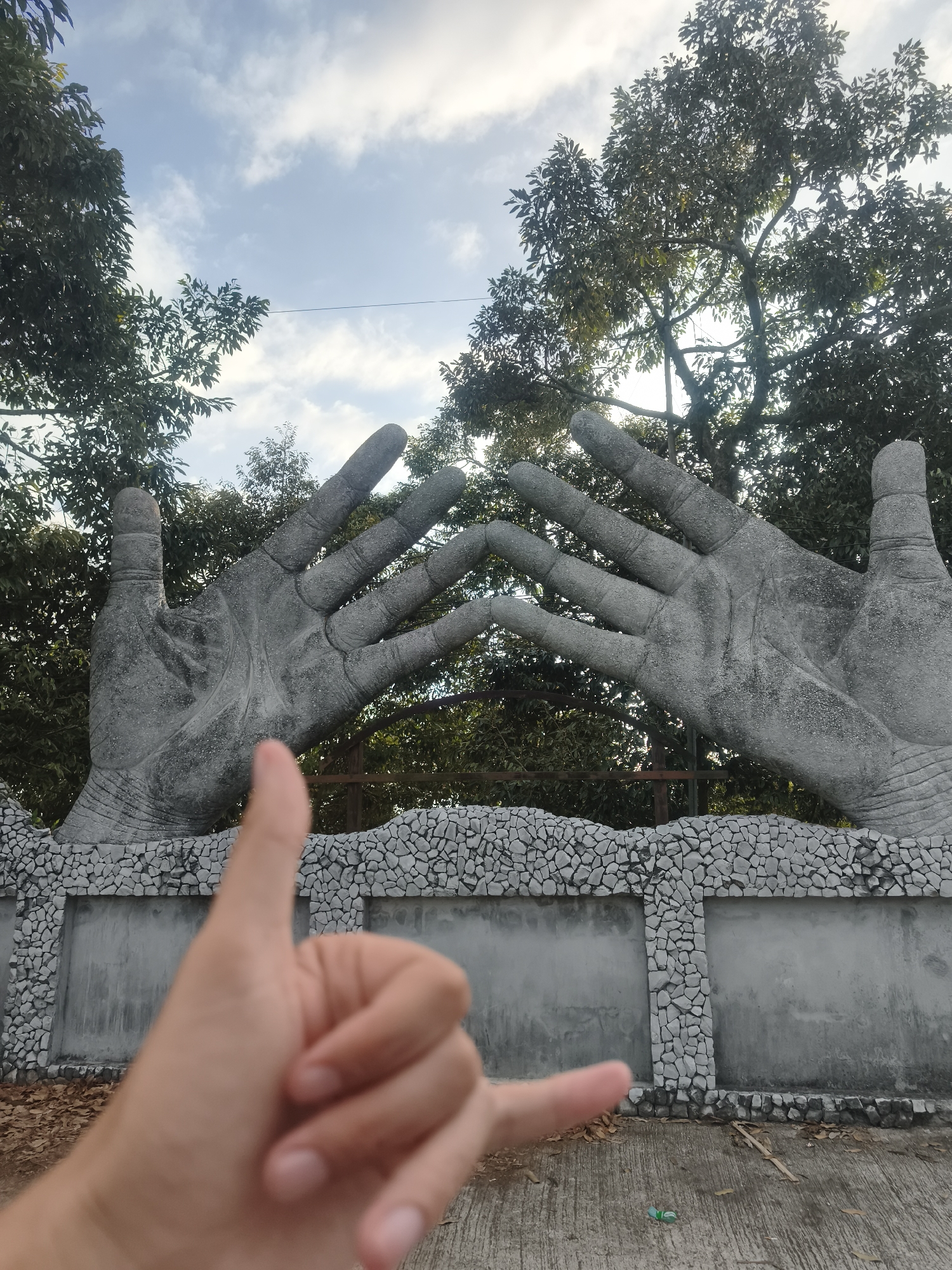 This is a photograph of a large sculpture of two hands with fingers extended, positioned on either side of a stone wall in what appears to be an outdoor public space in Ko Samui District, Thailand. The hands are made of stone and are detailed with wrinkles and fingernails. The wall below is decorated with a mosaic of natural stones. In the foreground, a person's hand is blurred, making a "shaka" hand gesture with their thumb and pinky extended. Behind the sculpture, lush green trees and a cloudy blue sky are visible. The ground in front of the wall is paved and appears to be an outdoor pathway or seating area. A single green bottle lies on the ground near the bottom right of the frame.
