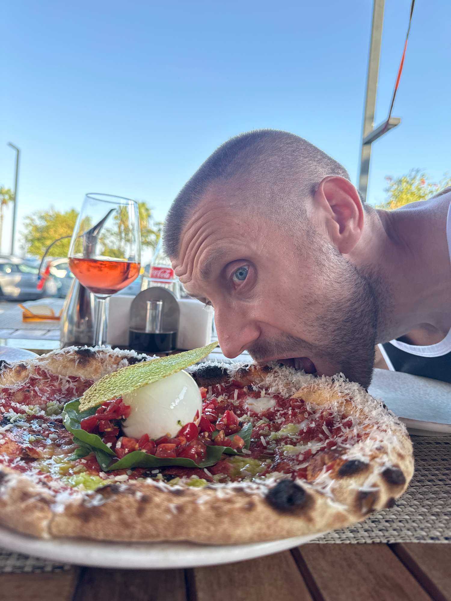 A man is leaning towards a pizza, with his mouth open as if to take a bite, in an outdoor restaurant setting. The pizza is topped with a ball of white cheese, chopped tomatoes, basil leaves, and a green garnish. A glass of rosé wine is visible in the background, along with a salt and pepper shaker, and a Coca-Cola bottle. The man has short hair and is wearing a white tank top. The environment appears to be a sunny day, with palm trees and a clear blue sky visible in the background, suggesting a Mediterranean or similar climate, possibly in Konyaaltı, Turkey, given the presence of palm trees and the overall ambiance often found in coastal resort towns. The table surface is a woven mat.