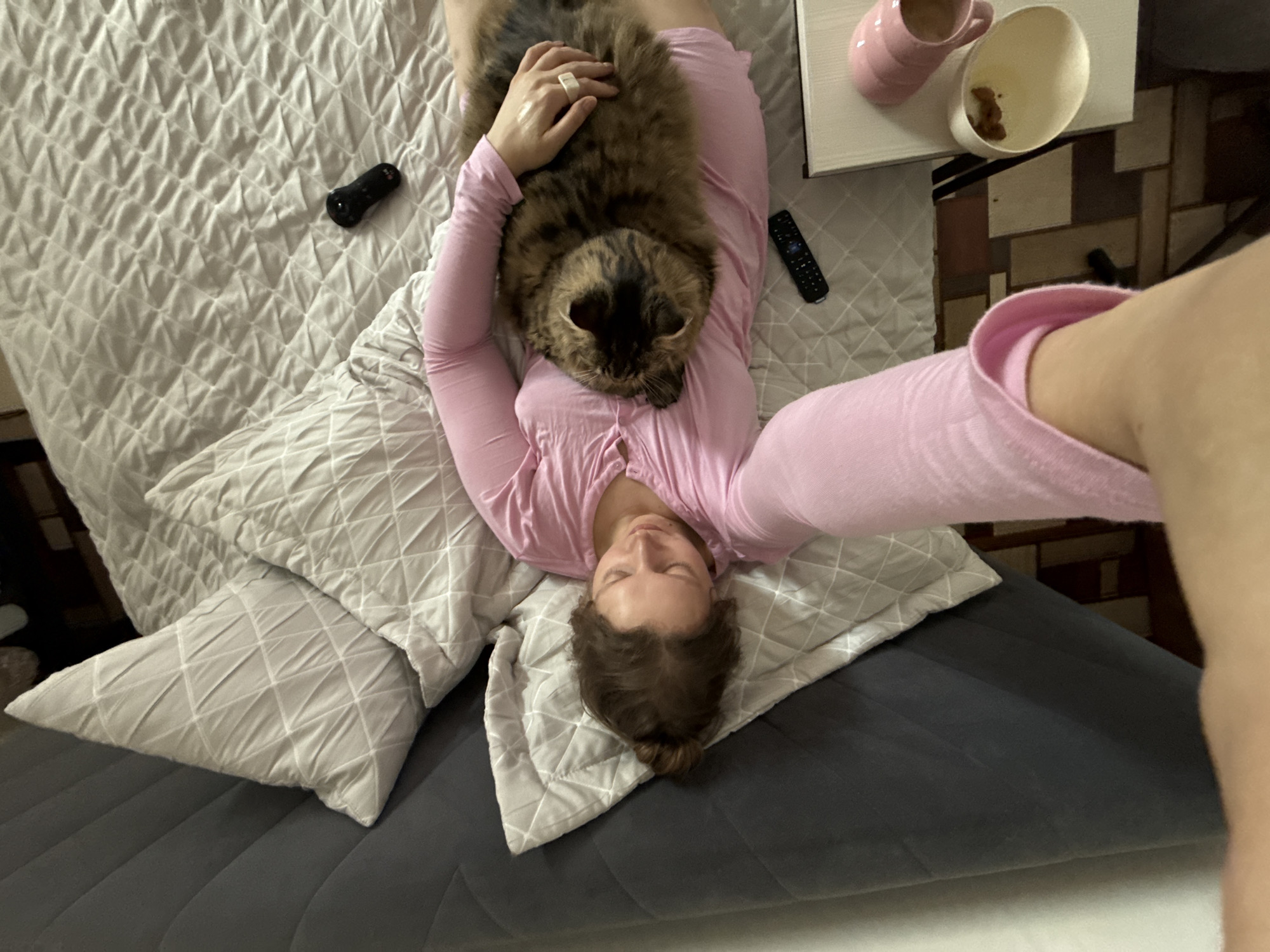 A woman in a pink long-sleeved shirt is lying on a bed with a gray comforter and white pillows. A tabby cat is lying on her chest. A remote control and a small black object are on the bed near her. A white bedside table holds a pink mug and a white bowl containing what appears to be crumbs. The background features what looks like flooring in a pattern of light and dark squares. The specific location within Izhevsk, Russia, cannot be determined from the visual information.