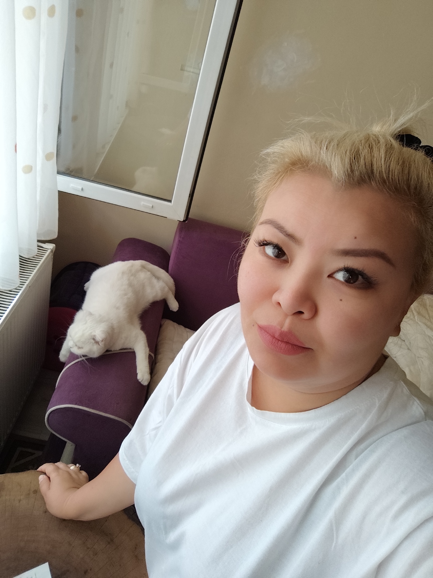 An indoor, slightly elevated selfie captures a woman and a white cat in a relaxed setting. The woman, positioned in the foreground, is looking directly at the camera with a neutral expression. She has blonde hair tied back, wears a simple white t-shirt, and has makeup including eyeliner and pinkish lipstick. A small mole is visible on her left cheek, and a ring is on a finger of her left hand, which rests on a wooden surface, likely a table.
Behind the woman, to the left, a fluffy white cat is sprawled in a seemingly deep sleep or highly relaxed state on a purple sofa armrest or cushion. The cat's body is stretched out, with its head facing away from the camera.
The background includes a window with a white frame, through which natural light enters, indicating it is daytime. A sheer white curtain with light brown polka dots hangs beside the window, and a white radiator is visible below it. The room's decor includes the purple sofa and a light-colored wall. The scene evokes a quiet, comfortable domestic atmosphere, potentially in Sarıyer, Turkey. No visible text is present.