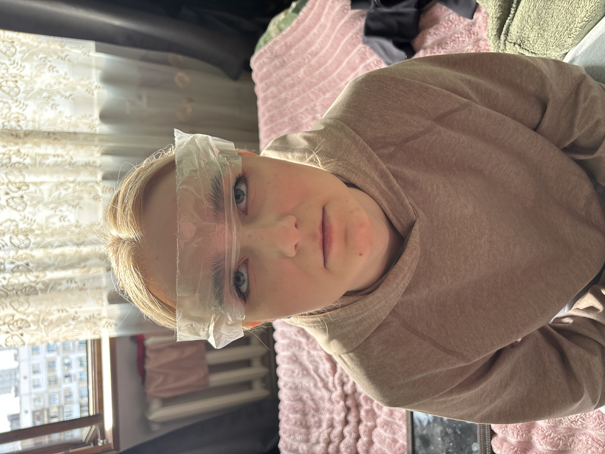 A young woman with blonde hair, wearing a tan hoodie, sits facing the camera with a serious expression. She has a piece of clear plastic wrap taped across her forehead, covering her eyebrows. The background suggests an indoor setting, possibly a bedroom. To her left, a window reveals a view of apartment buildings and what appears to be a radiator. To her right, a bed is covered with a pink textured blanket. The lighting indicates it is daytime, with sunlight filtering in from the window.