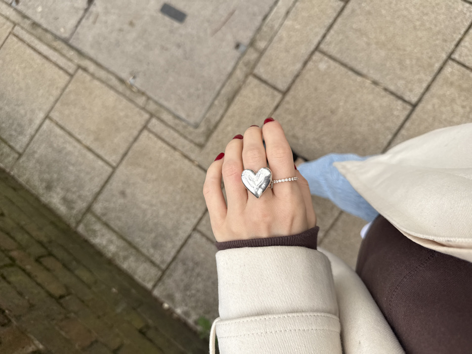 The image depicts an overhead perspective of an individual walking on a paved sidewalk in an urban setting. The primary subject is the person's left hand, adorned with dark red nail polish on the visible fingernails. A large, irregularly textured, silver-colored heart-shaped ring is worn on the ring finger, positioned above a slender band featuring a continuous row of small, clear stones. The individual is dressed in a light beige trench coat, layered over a dark brown garment, with light blue jeans visible on the lower body. A dark shoe is partially visible on the right foot, indicating forward movement. The ground is paved with light grey rectangular flagstones, showing signs of wear and surface dirt. To the left, a dark red brick wall with visible mortar lines exhibits areas of green discoloration, consistent with moss or algae. A dark, rectangular utility cover is partially visible within the flagstone paving towards the lower right. The architectural elements and paving style are consistent with a street environment in London, United Kingdom.