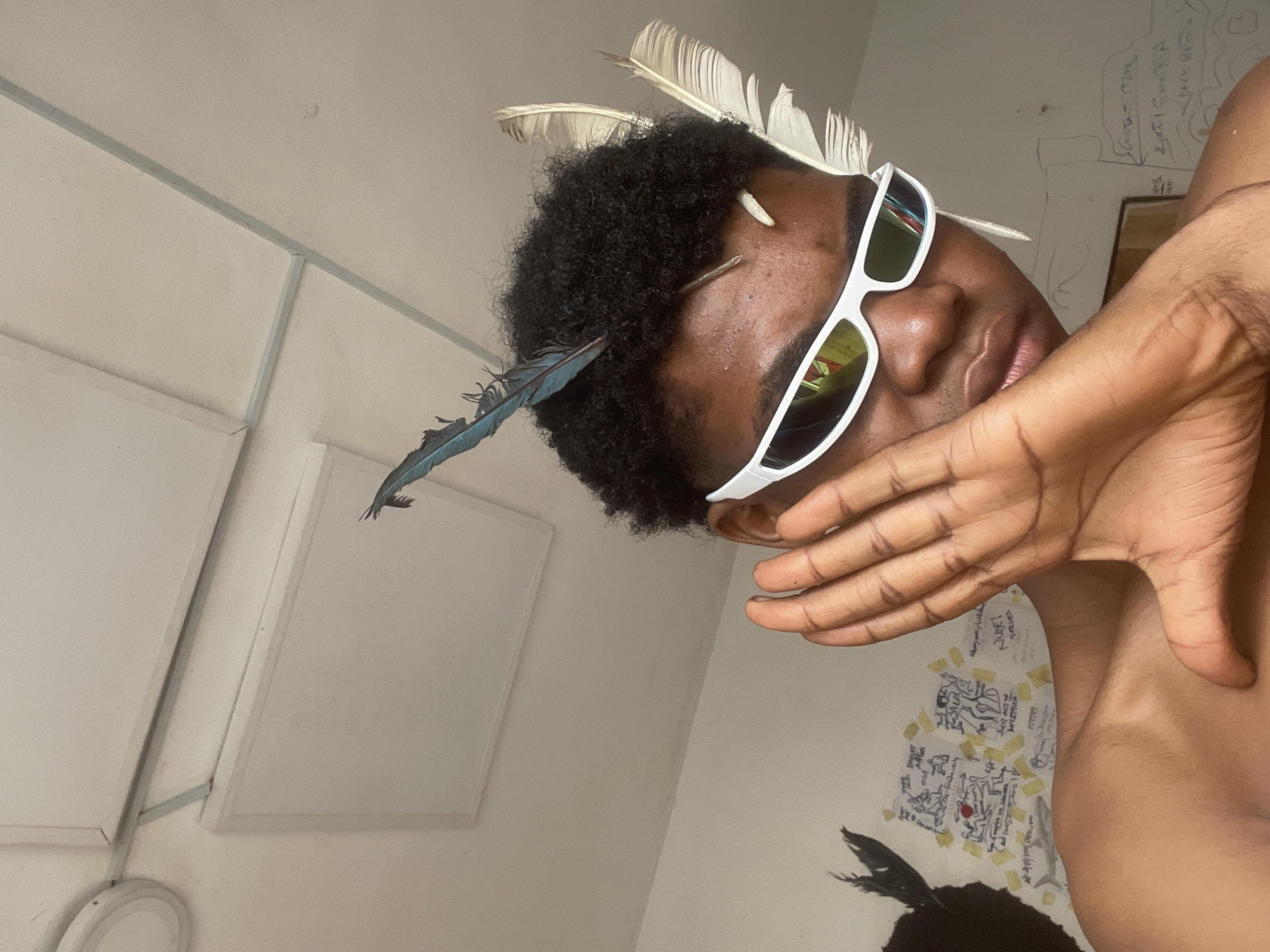 A person, wearing white futuristic sunglasses, is pictured from the chest up, in what appears to be a room with a white tiled ceiling. The person has dark, curly hair adorned with two blue feathers and two white feathers. Their right hand is raised to their face, obscuring their right cheek and eye. The background wall is white and features some handwritten notes and drawings. The lighting suggests an indoor environment. The scene is located in Ado, Nigeria.