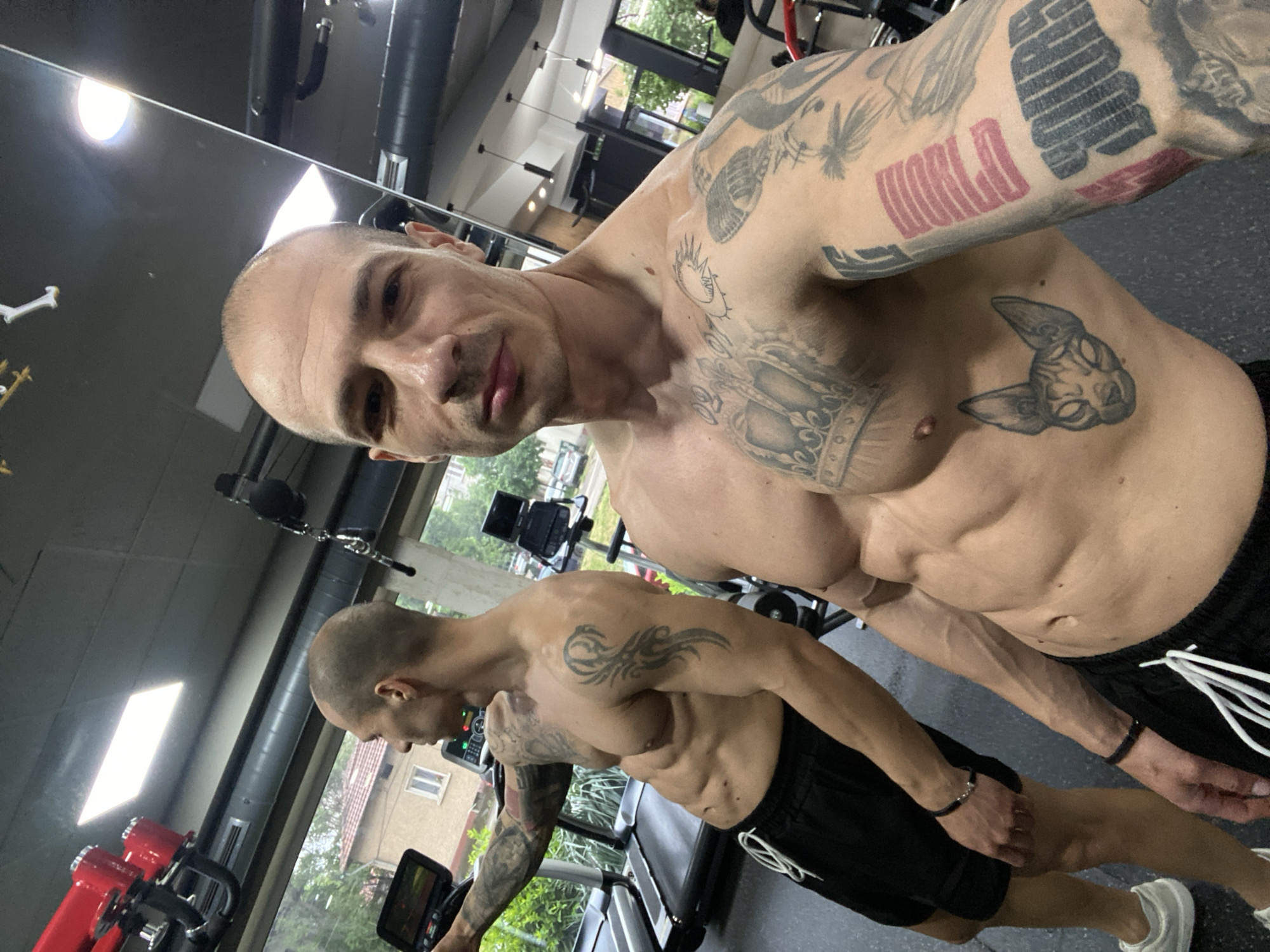 A shirtless, bald man with numerous tattoos is taking a selfie in what appears to be a gym in Pleven, Bulgaria. The man is fair-skinned and has a muscular physique. His tattoos include a design resembling a cat face on his chest, what appears to be a crown and additional designs on his right shoulder, and what may spell "WORLD" on his left arm. He is wearing black athletic shorts with white drawstrings and a dark bracelet on his left wrist.
Behind him and to the left, a treadmill is visible, along with another man who also appears to be shirtless and wearing black shorts. Gym equipment can be seen in the background, including various machines and weights. A window is visible behind the treadmill, offering a glimpse of buildings and greenery outside, suggestive of an urban environment.
The location is identified as Pleven, Bulgaria.
