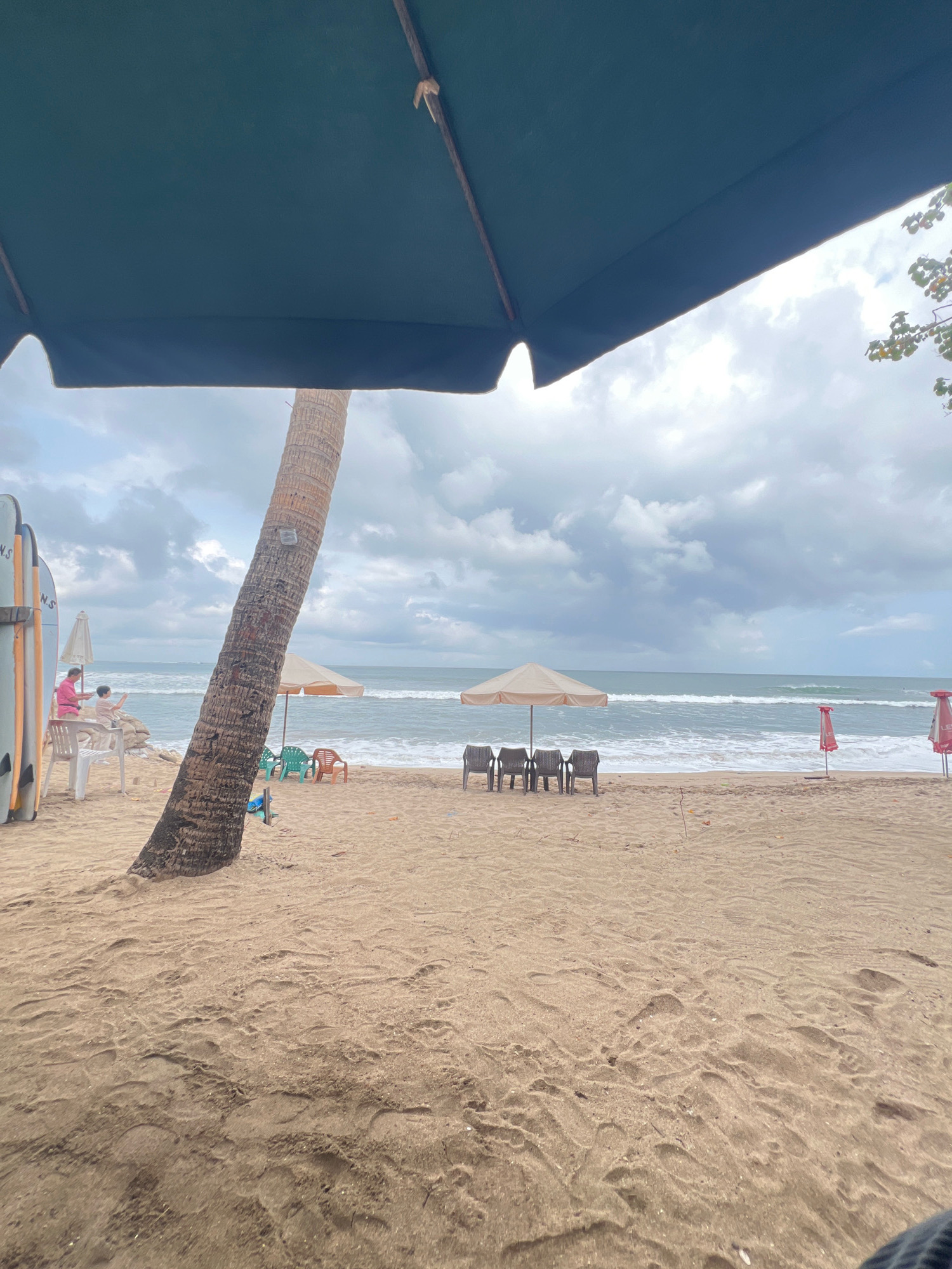 The image depicts a beach scene in Kuta, Indonesia. In the foreground, sand with footprints is visible, partially obscured by a large, dark green umbrella. To the left, a palm tree stands tall, leaning slightly towards the ocean. Several surfboards are propped up against a rack. Further back, on the sand, a person in a pink shirt and a child are near some white chairs and what appears to be a beige umbrella. In the middle ground, facing the ocean, are several chairs clustered under a beige umbrella. Two red umbrellas are positioned to the right. The ocean is visible in the background with waves rolling in under a cloudy sky.