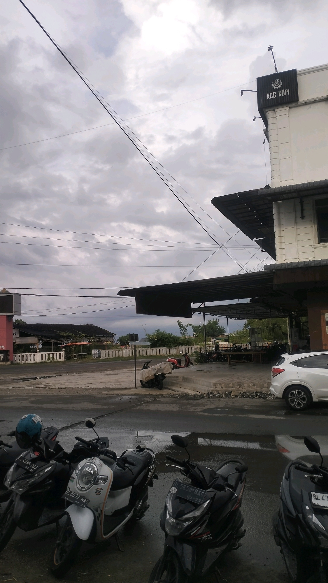 The image depicts a street scene in Lhokseumawe, Indonesia, under a cloudy sky. In the foreground, three motorcycles are parked on a wet asphalt surface. The middle motorcycle is white with black accents and license plate "BL 5682 NAG 09-23". To its left, a black motorcycle has a blue helmet on its seat and license plate "BL 4686 DAG 09-24". To the right of the white motorcycle, another black motorcycle has license plate "BL 5658 NAC 09-23".
Behind the motorcycles, a low-rise building with a white facade and a dark-tiled roof is visible. A sign with "ACC KOPI" in white letters on a black background is affixed to the upper corner of the building. A covered seating area, likely an outdoor cafe or restaurant, extends from the building, supported by dark metal poles. Tables and chairs are visible under this canopy. Further back, another building with a dark, sloped roof and pink accents is present. In the distance, a white car is parked. The ground is wet, suggesting recent rain, and reflects the cloudy sky and surrounding structures. Power lines crisscross the sky. The overall atmosphere is overcast and subdued.