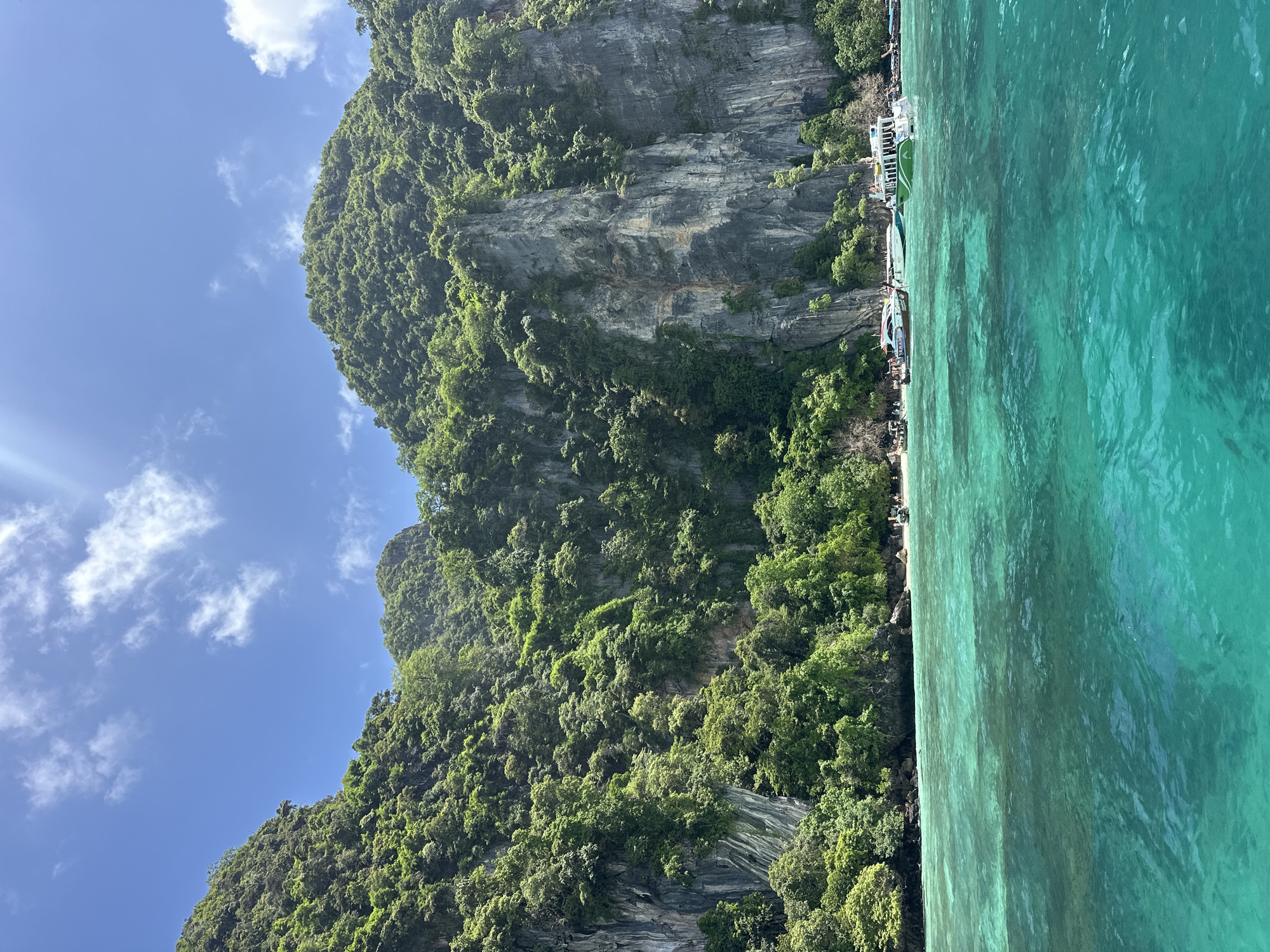 A wide-angle, eye-level shot captures a tropical scene of verdant, jungle-covered cliffs rising steeply from a turquoise sea. The cliffs are rugged and grey, with streaks of brown and white running down their faces. Lush green vegetation clings to the steep slopes and rocky outcrops. The water in the foreground is crystal clear, revealing the sandy bottom in shallower areas. Several boats are anchored near a small, sandy beach nestled at the base of the cliffs. People can be seen on the beach and on some of the boats. The sky above is a bright blue, dotted with fluffy white clouds. The overall impression is one of a serene and beautiful natural landscape. This scene is located in the city of Haad Hin Khom, Thailand.