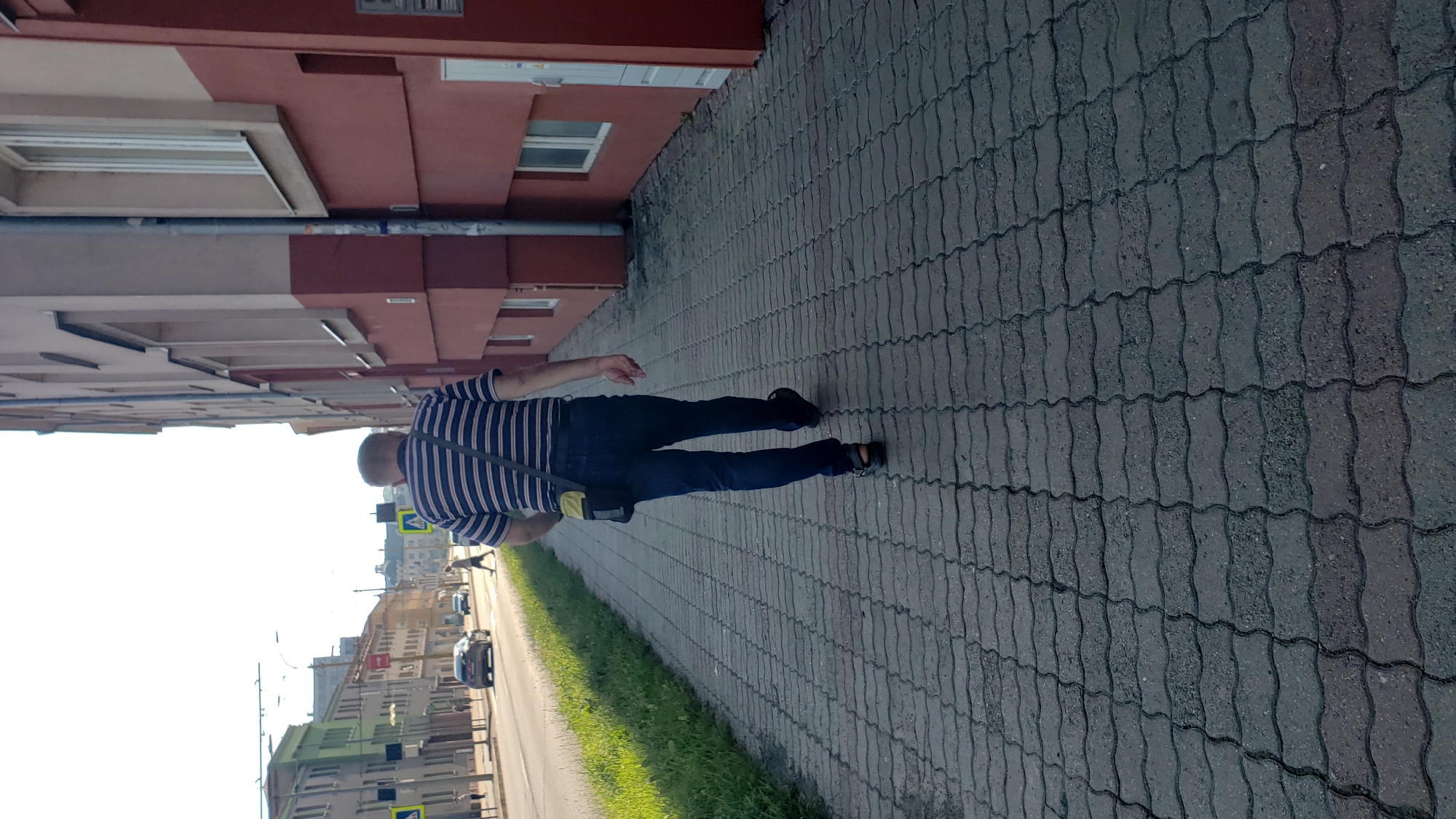 An individual is walking away from the camera on a paved sidewalk adjacent to a street with traffic. The person is wearing a striped t-shirt, dark blue jeans, and sandals, with a small bag slung across their body. Behind the sidewalk is a grassy area and a red-colored building with windows. Across the street, buildings with green and other facades are visible. The scene appears to be in an urban setting, likely in the city of Prešov, Slovakia, given the architectural style and street layout. The sky is bright and appears to be daytime, with the sun casting shadows.