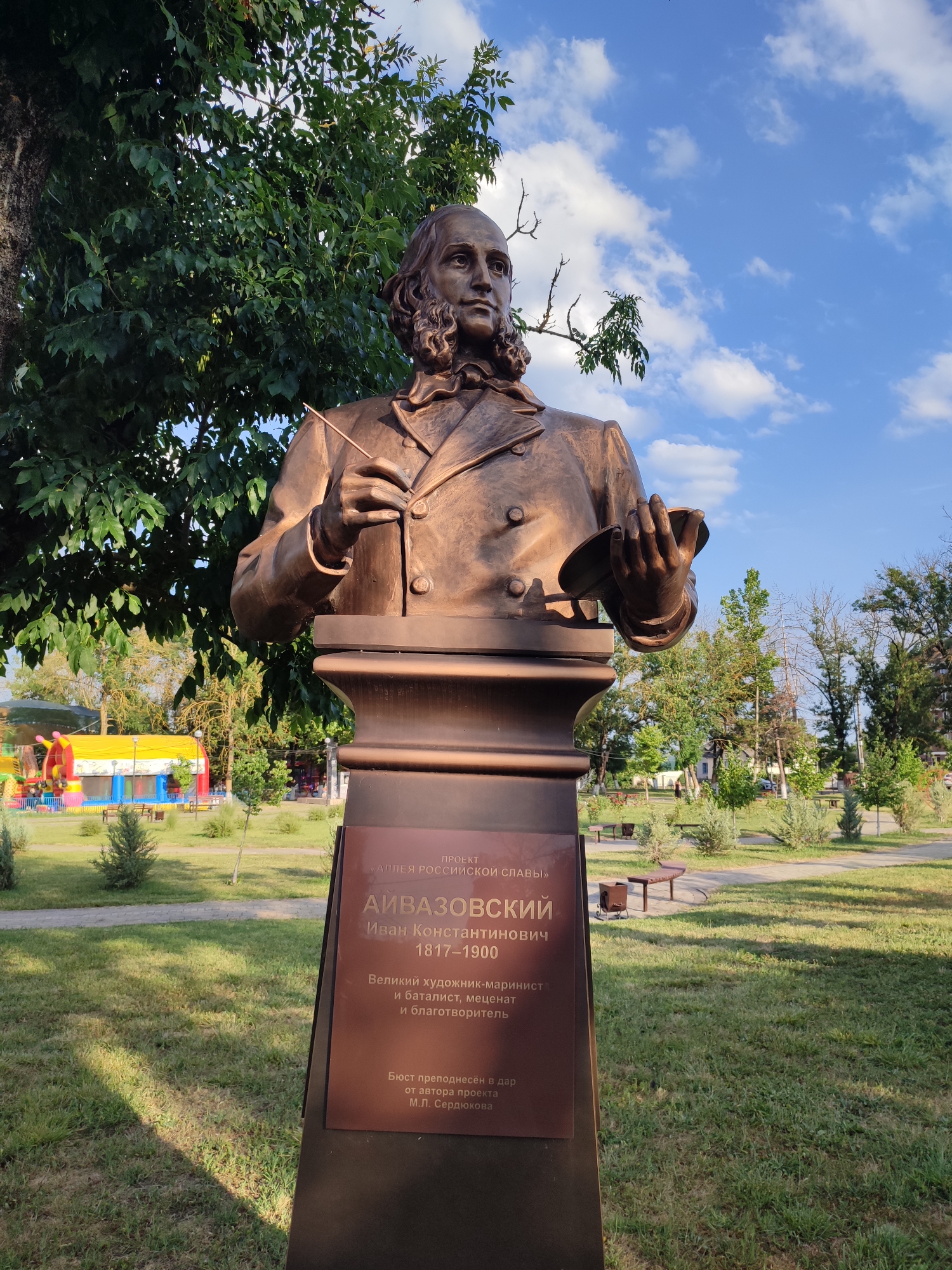 A bronze bust of a man, identified by a plaque as Ivan Konstantinovich Aivazovsky (1817-1906), stands on a pedestal in an outdoor setting. The bust depicts the man with his hair tied back, wearing a formal jacket with a ruffled collar. He holds a paintbrush in his right hand and a palette in his left. The plaque below the bust states it is part of the "Alley of Russian Glory" and describes Aivazovsky as a "Great artist-marinista, marine painter, philanthropist and benefactor." The bust is located in Abinsk, Russia, as indicated by the context of the "Alley of Russian Glory." The background shows a park-like environment with green grass, trees, benches, and a colorful playground structure in the distance. The sky is blue with scattered clouds.