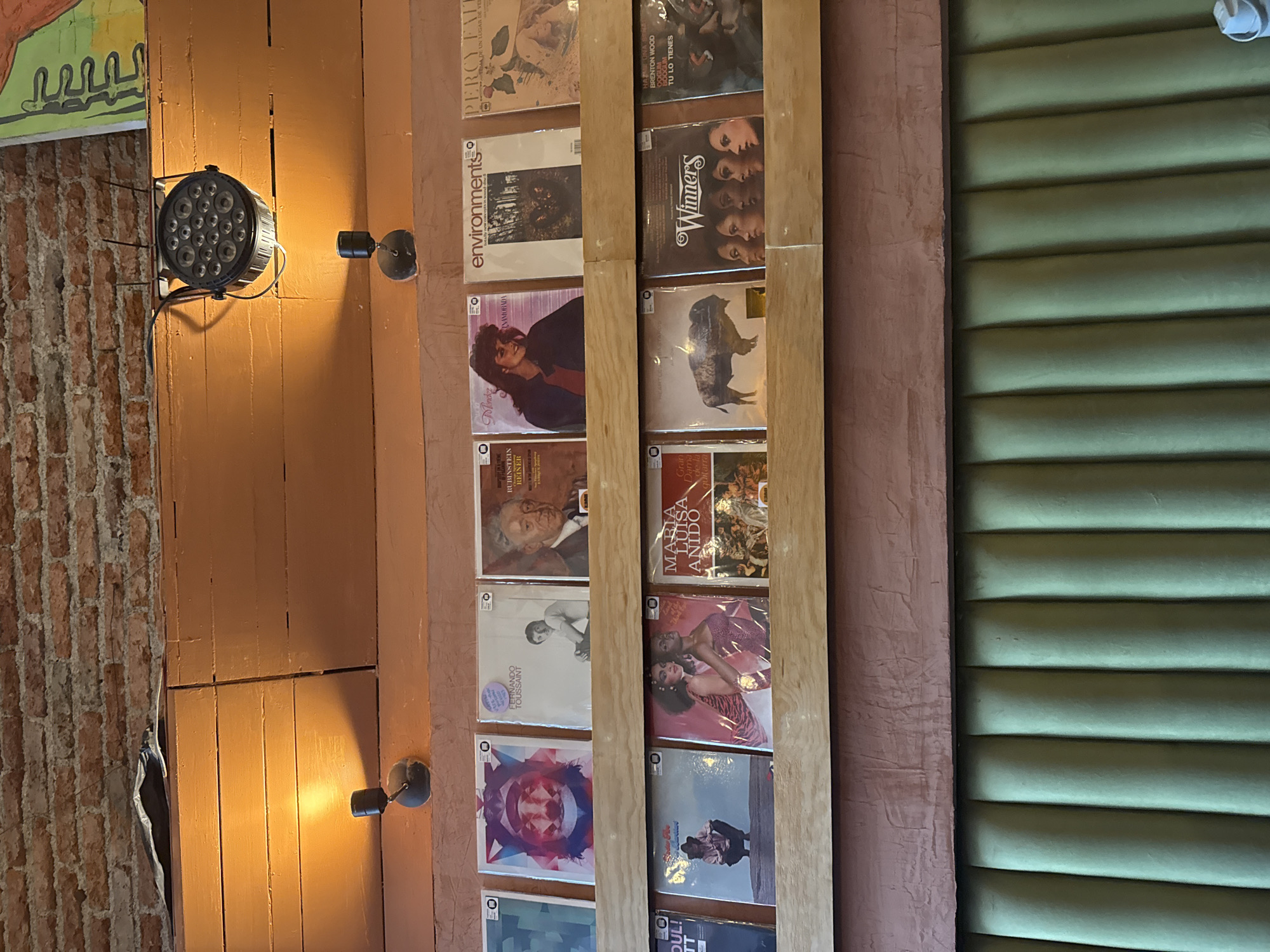 Here is a description of the image.
The photograph shows a vertical display of what appears to be vintage record albums mounted on a wooden frame affixed to a wall. The wall, possibly wood paneling, is painted in warm tones. To the left of the album display is a brick wall section and a wall light fixture. The right side of the image shows a panel with horizontal slats, potentially a window covering or decorative element.
Based on the text snippets visible in the image, like "Winner's", "Environments," and names of musicians, it's plausible the albums are music records. The lighting in the photograph suggests an indoor setting, possibly a music store, a bar, or a private collection.