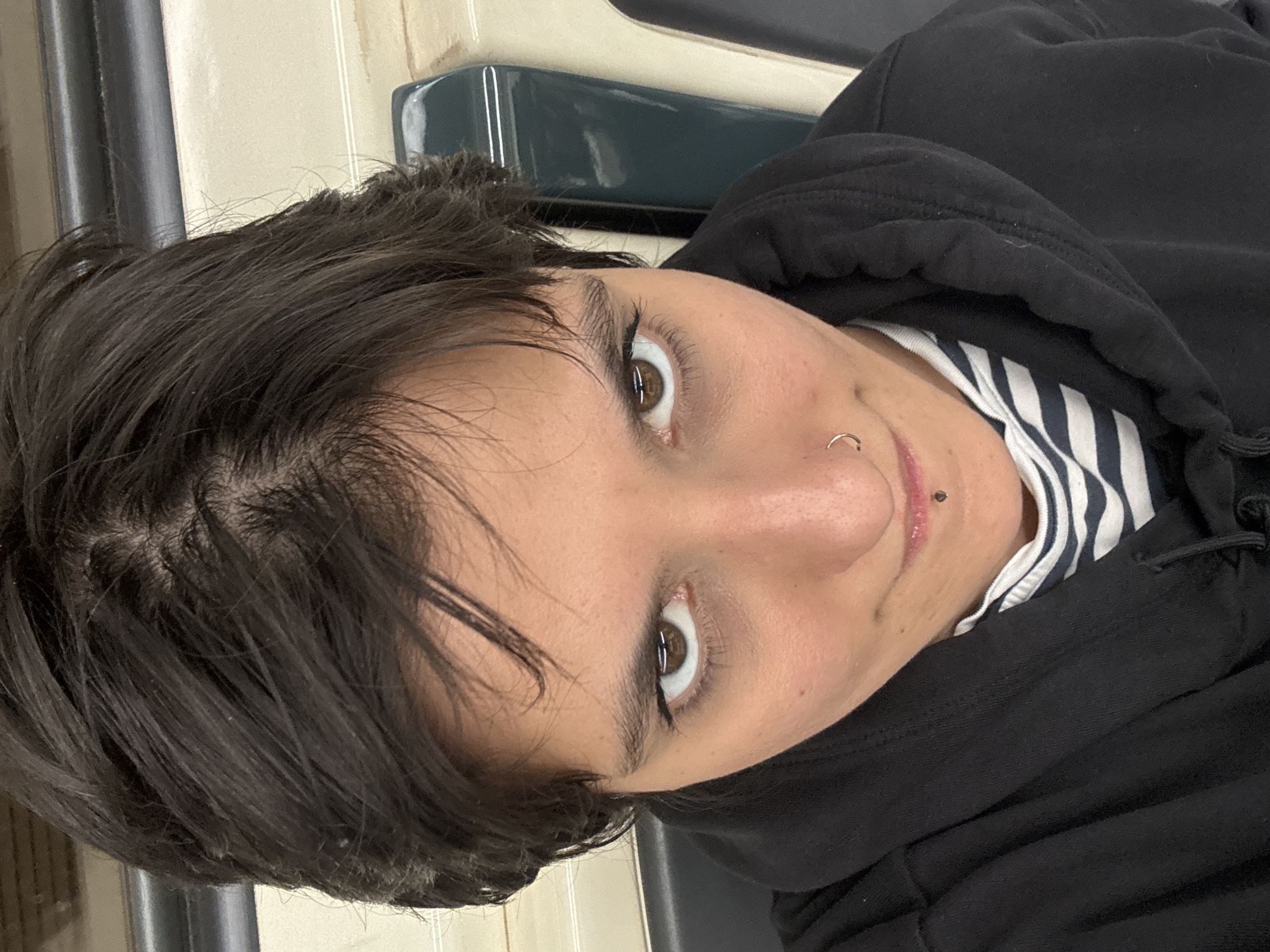 A close-up, upward-angled photograph depicts a person indoors, possibly on public transportation. The person has dark hair, light skin, and brown eyes. They are wearing a black hooded sweatshirt over a black and white striped shirt. A nose ring is visible in their left nostril and a labret piercing is seen below their lower lip on the right side. The background includes a dark green surface and a cream colored wall with gray or silver vertical stripes. The location is potentially the Sofia, Bulgaria subway system, suggested by the seating design.