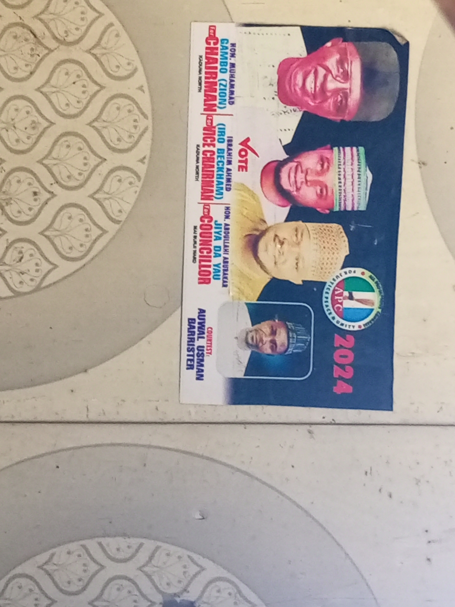 A campaign poster for the All Progressives Congress (APC) party for the 2024 election is shown on a patterned tile surface. The poster features images and names of four candidates, including Hon. Muhammad Gambo (Zion) for Chairman, Ibrahim Ahmed (Iro Beckham) for Vice Chairman, Hon. Abdullahi Abubakar Jiya Da Yau for Councillor, and an individual whose name is Auwal Usman Barrister. The location information "Mai Burji Ward" indicates the specific geographic area in Maiburiji, Nigeria, where the candidate for Councillor is running. The poster includes the APC logo and the word "VOTE" with a check mark.