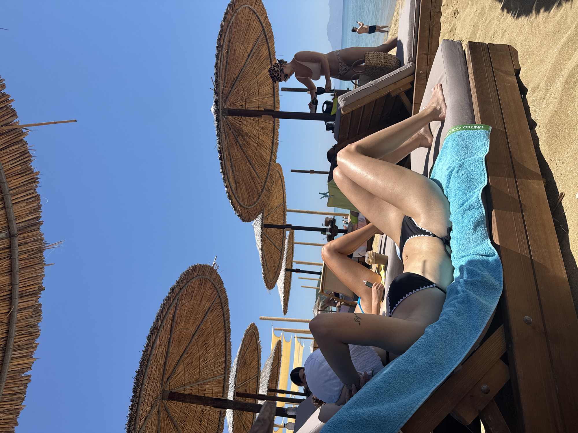 The photograph depicts a beach scene in Nea Peramos, Greece. The camera is positioned at ground level, looking up towards the sky, which is a clear, vibrant blue. Several thatched beach umbrellas line the sand. A woman in a black and white bikini is reclining on a beach chair, covered by a light blue towel. Another woman in a beige bikini, headscarf, and sunglasses stands at a beach chair nearby, placing drinks on a tray. In the background, near the sea, a person is partially submerged in the water. The beach is sandy and appears to be well-maintained, with other beachgoers visible in the distance.