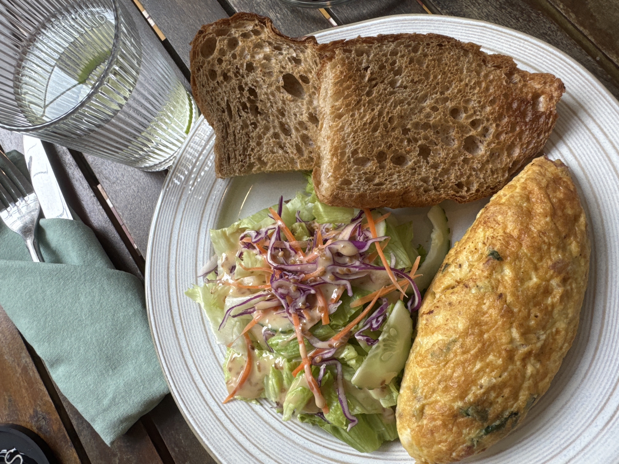 On a wooden table sits a plate holding a meal consisting of two slices of toasted bread, a green salad with shredded purple cabbage and carrot, cucumber slices, and a creamy dressing, as well as an omelet. A glass of water with a lime slice is visible next to the plate, accompanied by silverware and a light green napkin. A partially visible black object is in the bottom left corner. The location cannot be determined.