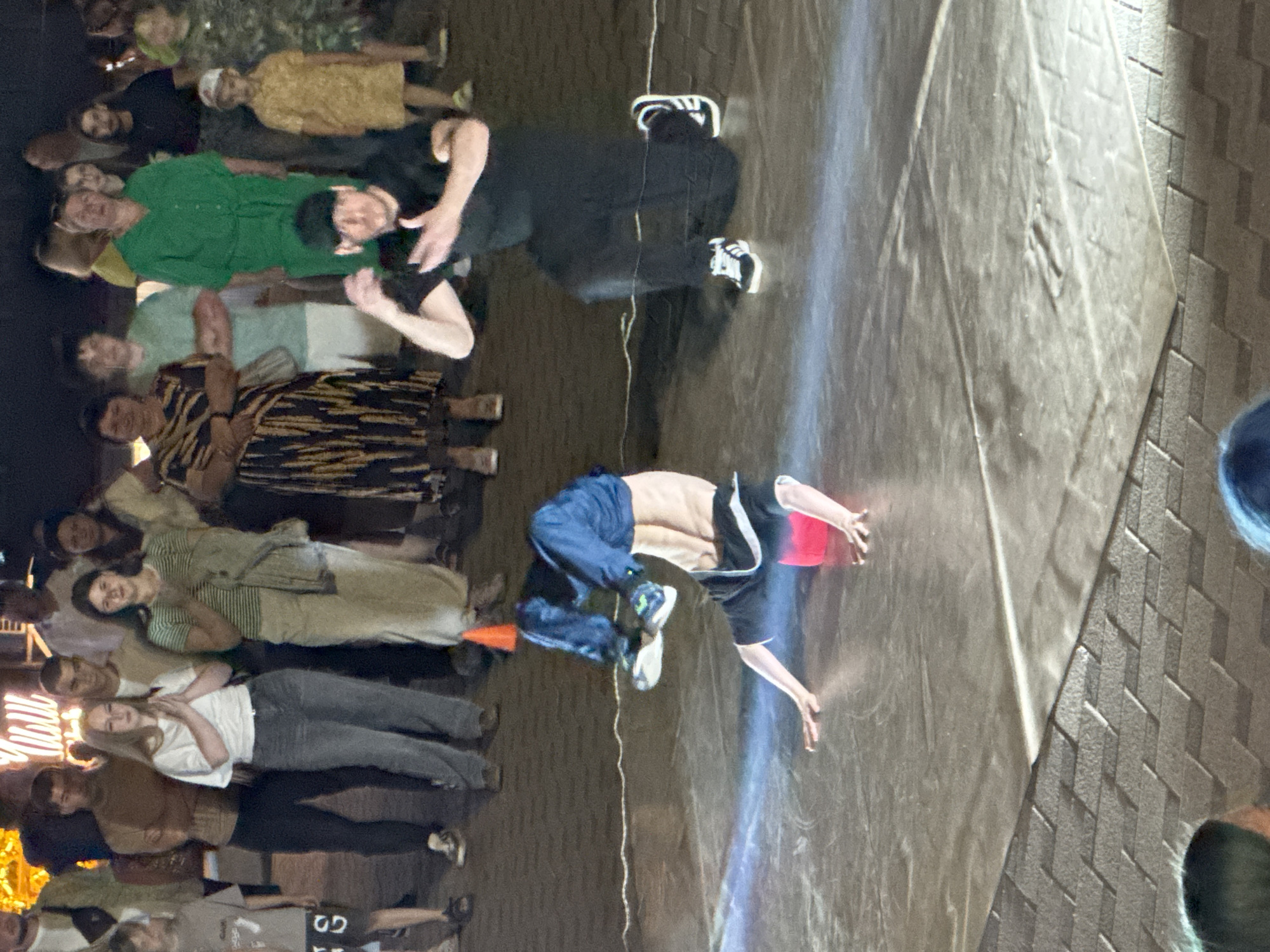 In the city of Unknown, Unknown, a group of people are gathered outdoors at night to watch a breakdancer perform. The breakdancer is in the foreground, on their head with their legs in the air, wearing a blue shirt and black shorts. Another man, wearing a black t-shirt and black pants, is kneeling on the ground in front of the breakdancer, appearing to be part of the performance or a fellow dancer. A crowd of spectators, including men, women, and children, are standing behind the performers, observing the show. The ground appears to be paved with cobblestones. There are some cones on the ground, and a spotlight illuminates the breakdancer. The overall scene suggests an informal outdoor performance or event.