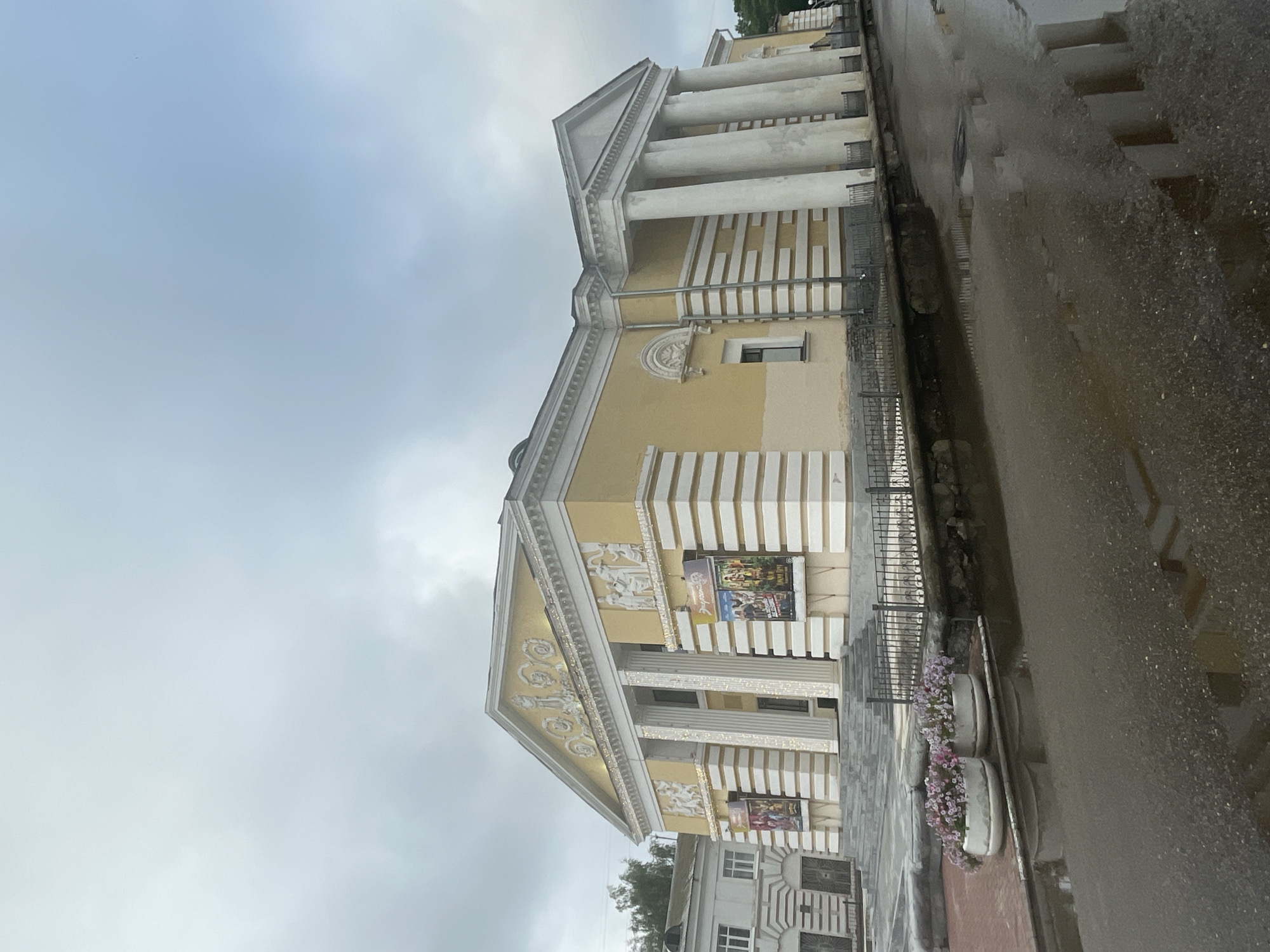 The image shows the Zavolzhsky City Drama Theater in Zavolzhye, Russia, on an overcast day. The theater is a grand, neoclassical building with a yellow facade, white columns, and decorative friezes. A wide staircase leads up to the entrance. In front of the theater, there is a black metal fence bordering a wet street with puddles reflecting the building and the sky. Two white planters filled with pink and purple flowers are situated on the sidewalk to the left of the stairs. Posters are displayed on the walls of the theater, indicating current performances or attractions. The overall scene suggests a somewhat damp and quiet atmosphere, characteristic of a cloudy day in a city square.